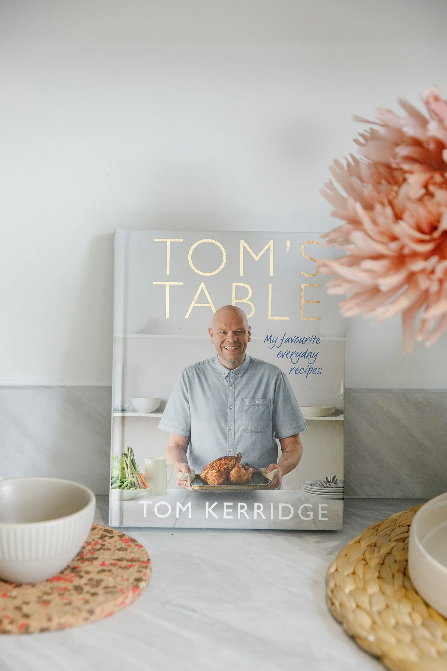 A book cover featuring a man in a light blue shirt holding a cooked turkey, with vegetables and a pitcher on the table. The title is "Tom's Table" by Tom Kerridge, with a large pink flower and a bowl in the foreground.