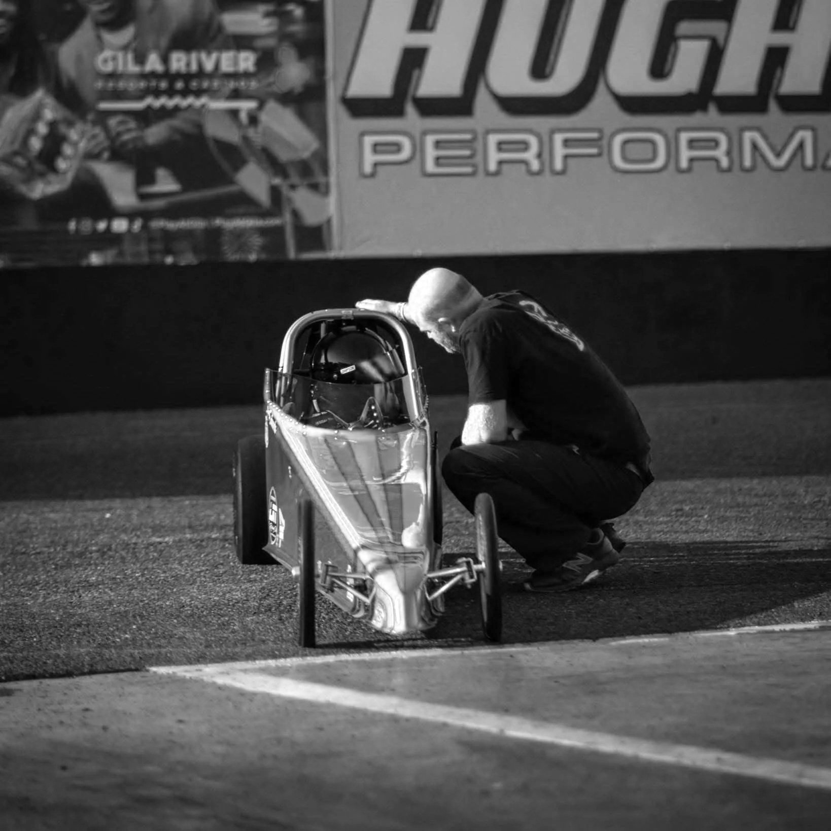 A person kneeling beside a drag race car on a track at night, inspecting or working on the vehicle.