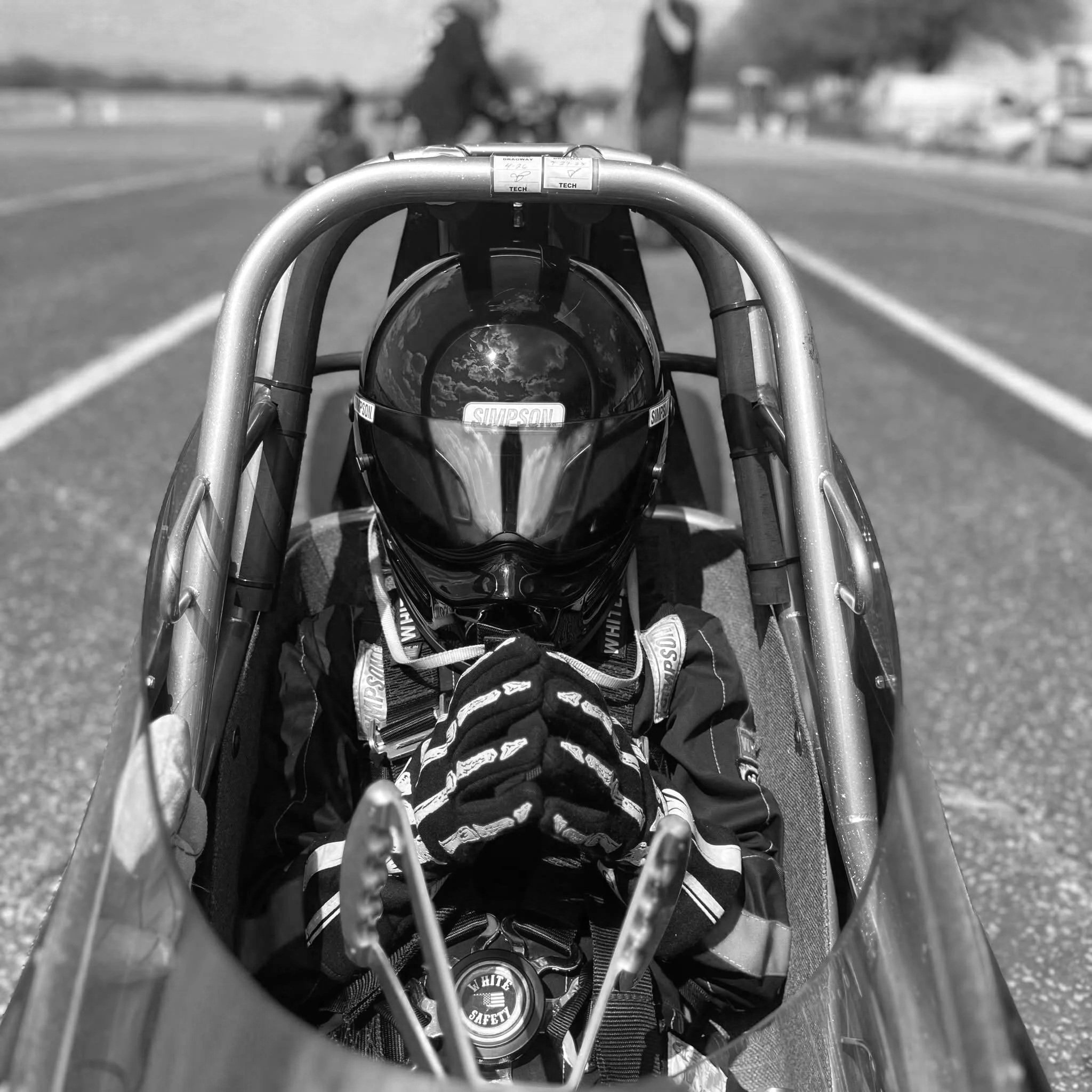 Black and white photo of a person in a racing helmet and gloves inside a race car, with their hands clasped in front of their face, on a racetrack.