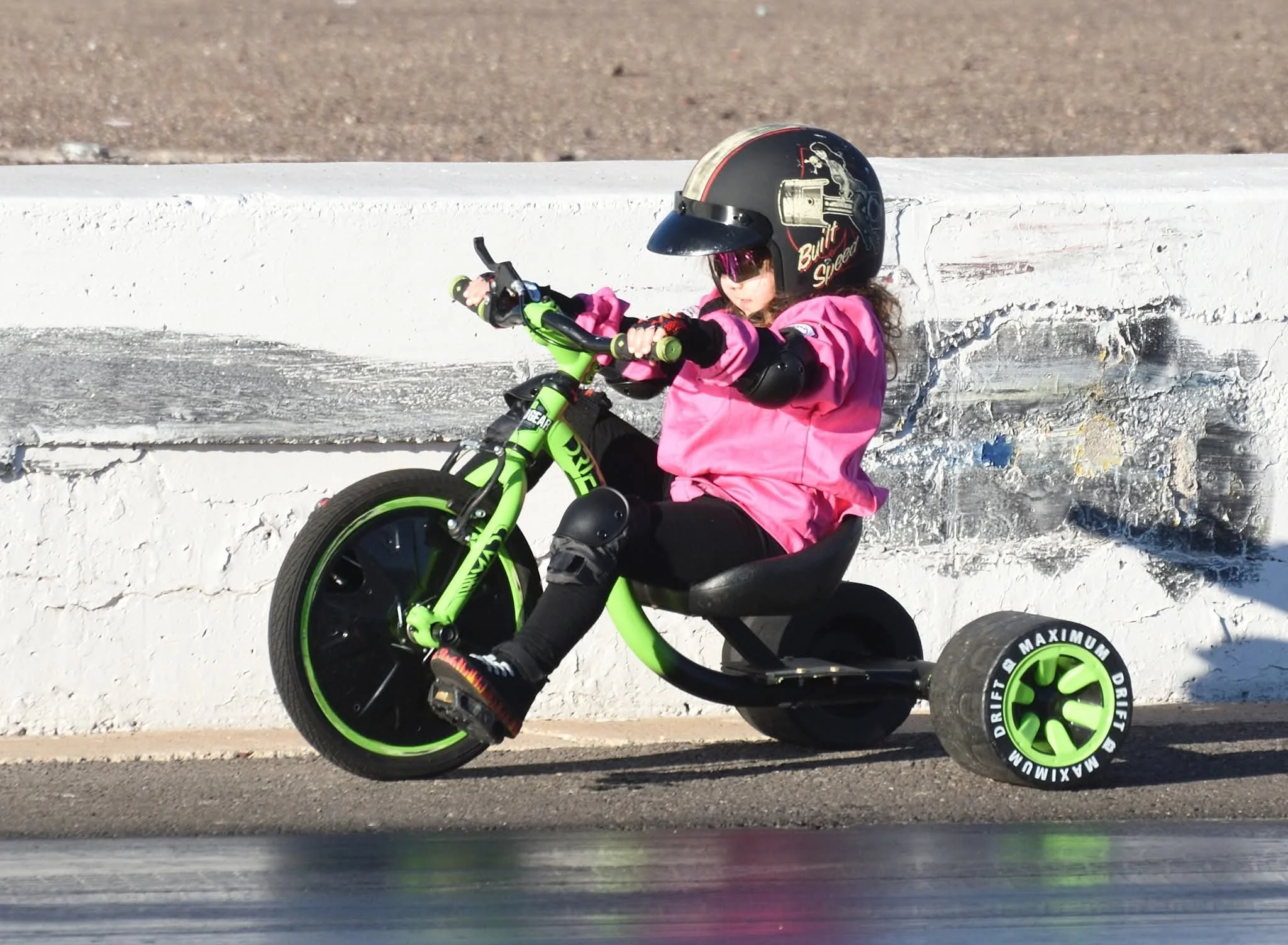 A young girl wearing a black helmet, pink jacket, and sunglasses riding a bright green and black small bicycle with training wheels along a street, with a white textured wall and concrete curb in the background.