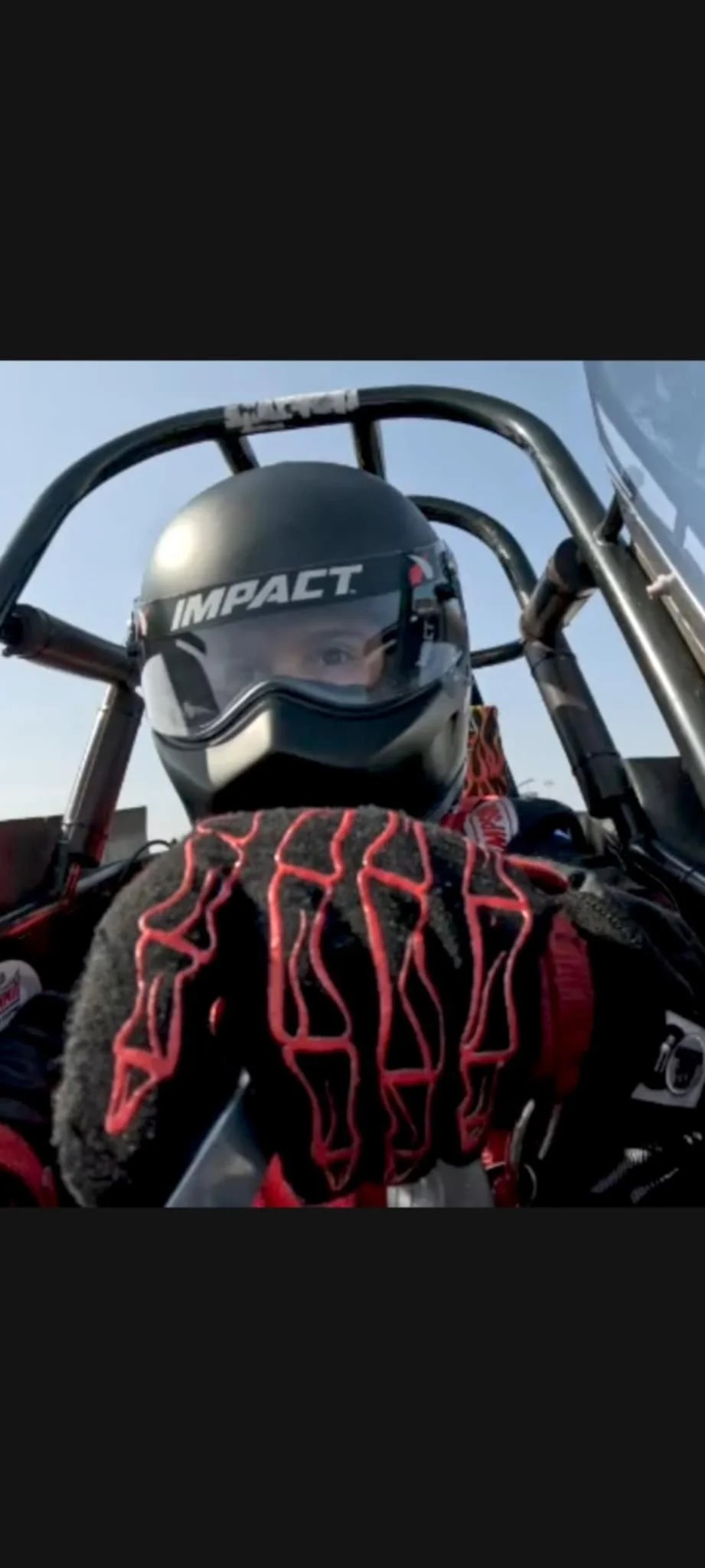 Close-up of a race car driver in a helmet and racing suit, sitting in a racing vehicle with a clear sky background.
