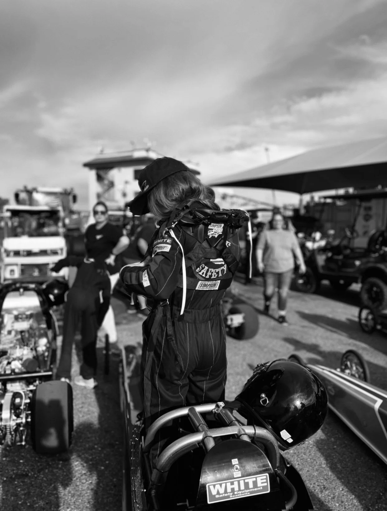 A woman in racing gear adjusting her helmet on a go-kart in a paddock area with other people, tents, and equipment around.