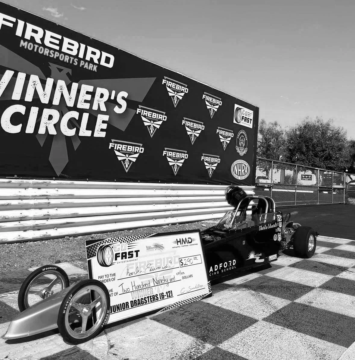 A drag racing car and oversized check displayed at Firebird Motorsports Park, with a backdrop of banners including Firebird logos and NHRA branding. The check is made out to Charlie Edwards for $290, awarded for winning a Junior Dragsters (6-12) race.