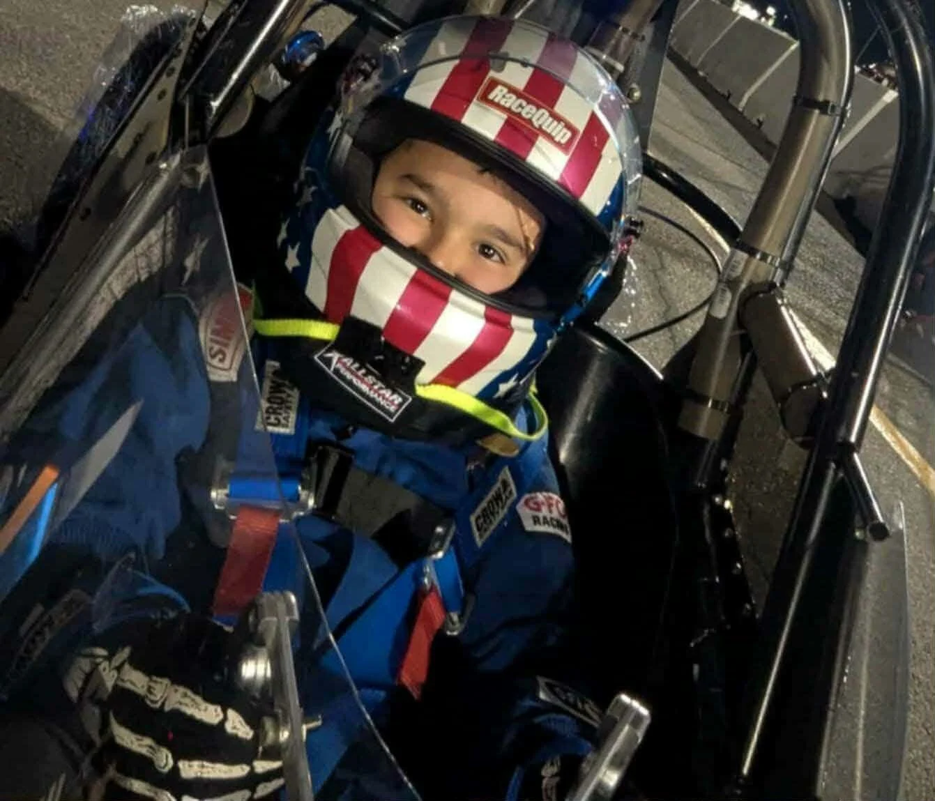 A young child in a racing suit and helmet sitting in a go-kart, smiling at the camera. The helmet has red, white, and blue stripes, and the racing suit has various sponsor patches. The go-kart has a transparent windshield and visible metal frame components.