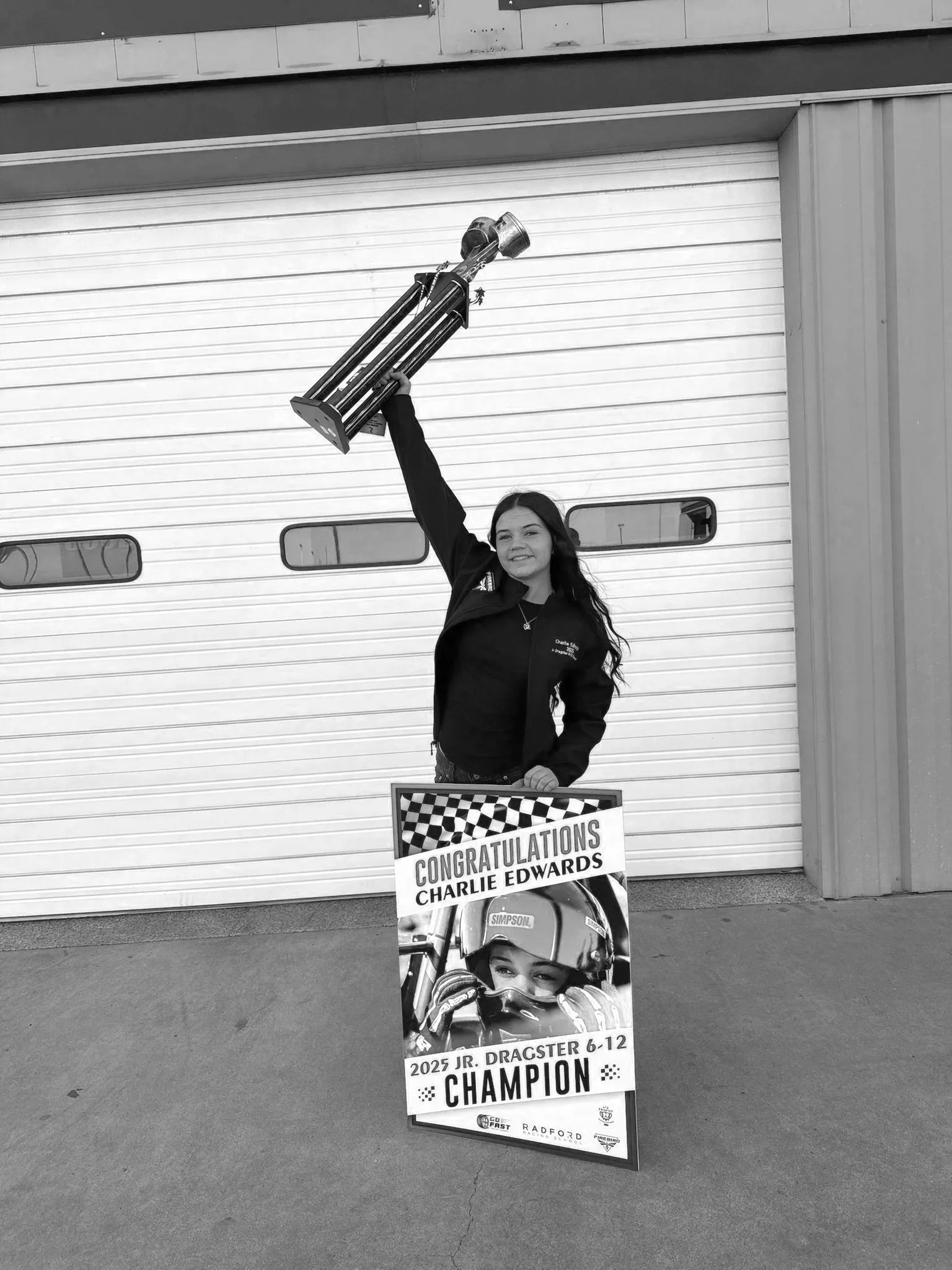 Young girl holding a trophy in one hand and a championship sign with a racing driver on it in the other, standing in front of a garage door. The sign congratulates Charlie Edwards as the 2025 Jr. Dragster champion for ages 6-12.