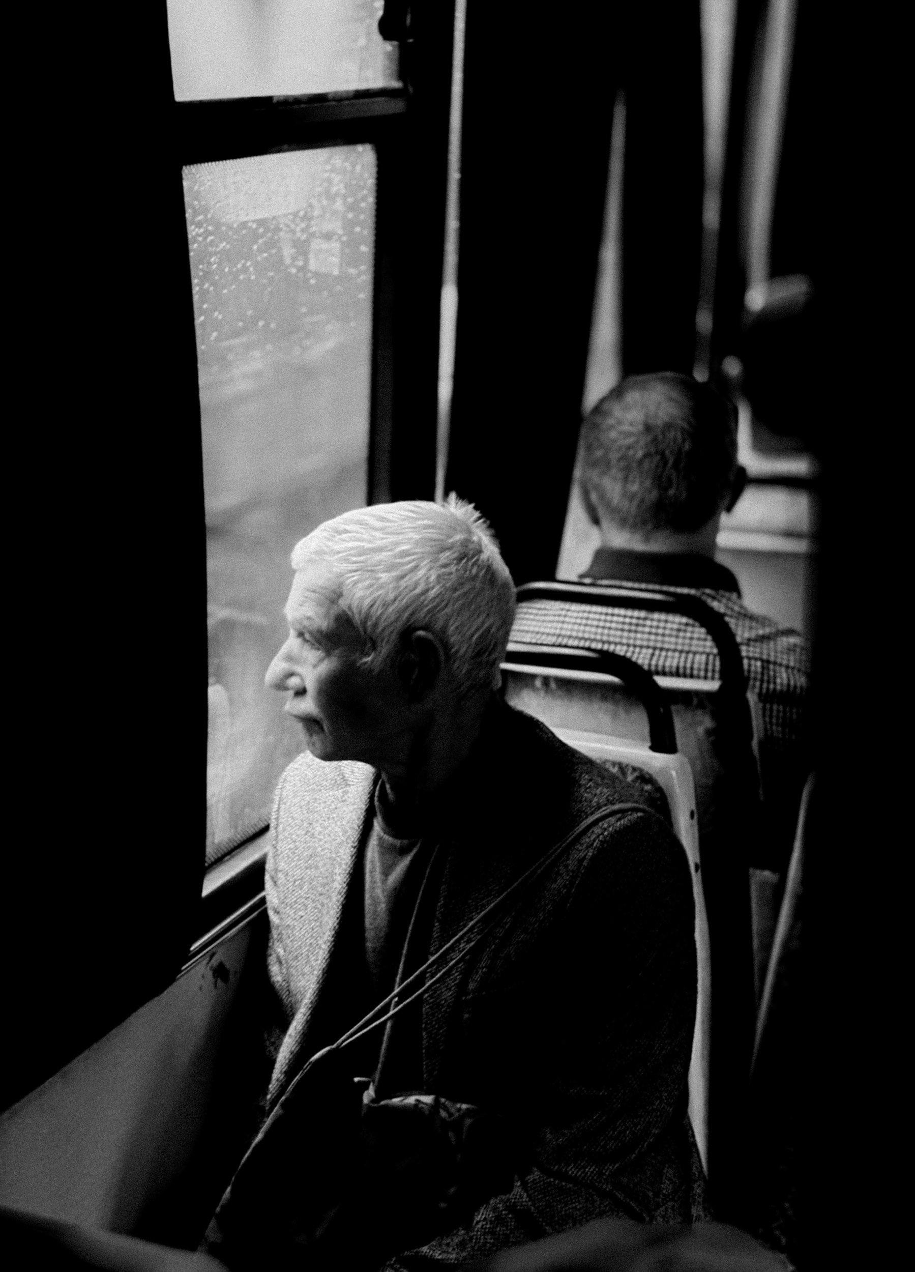 An elderly man with white hair and glasses, sitting on a bus, looking out the window at traffic and buildings. He is wearing a jacket and has a bag or camera strap across his shoulder. The photo is in black and white.