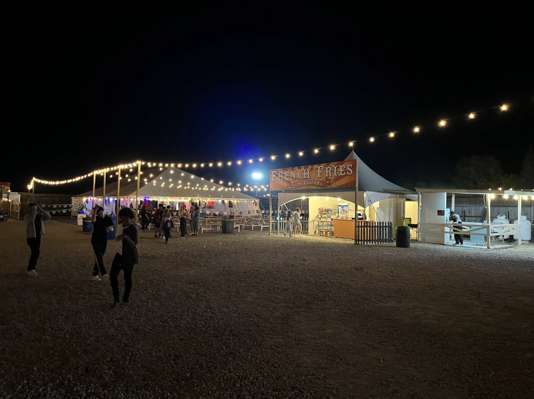 Night scene at a fair with string lights, a food stand labeled 'French Fries Concessions,' and people walking around.