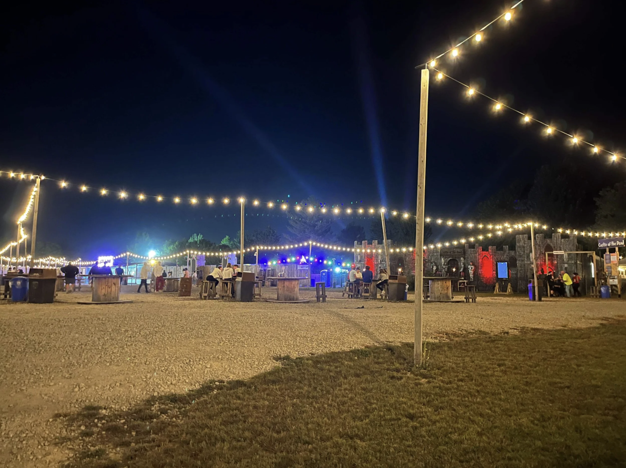 Night scene of an outdoor fair or carnival with string lights overhead, people sitting at tables, and a castle amusement ride in the background.