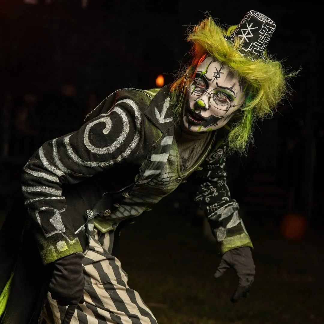 Person dressed as a clown with black and white face paint, green and yellow hair, and wearing a patterned jacket, posing in a dark setting.