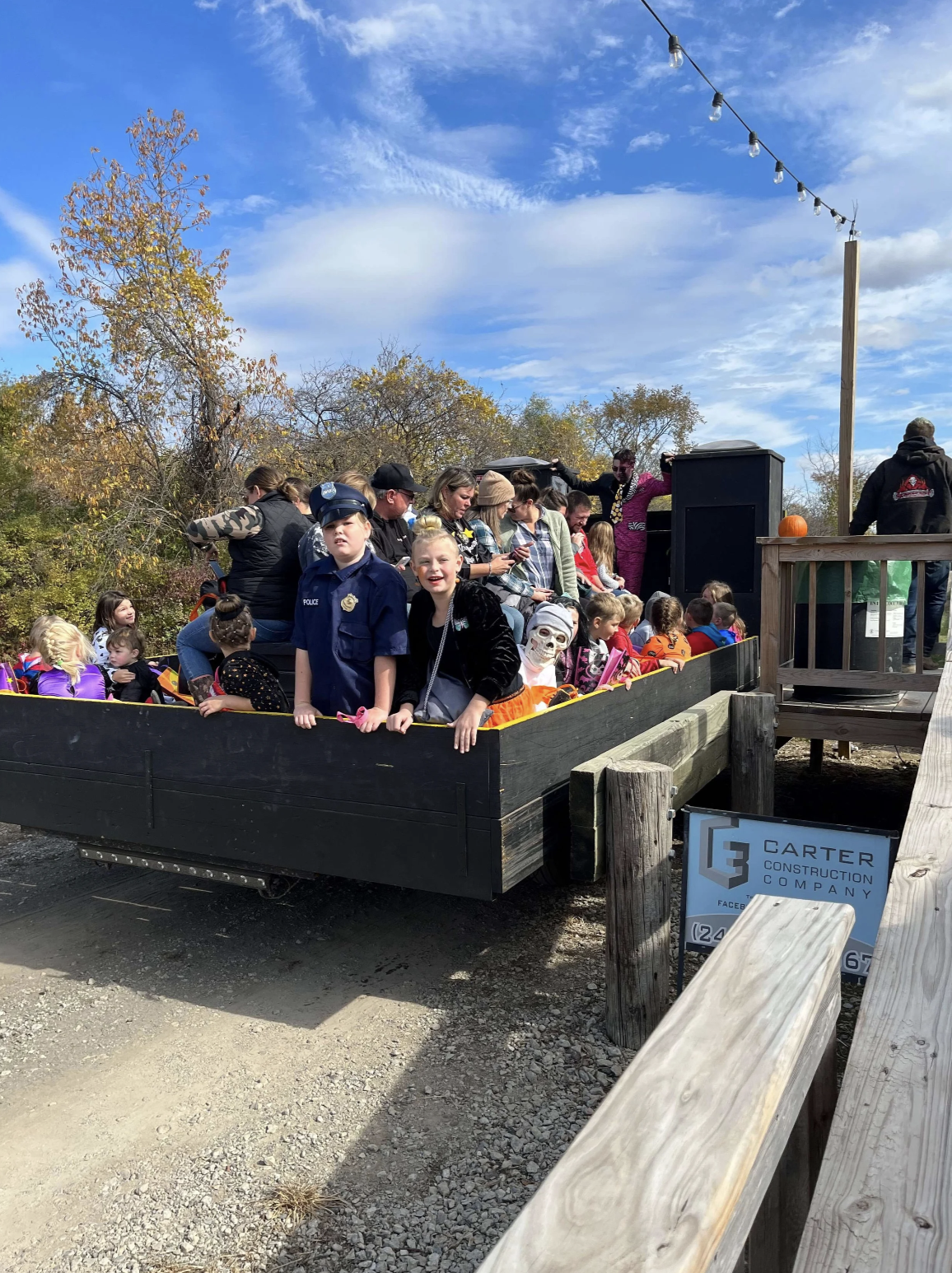 Children and adults in Halloween costumes riding a haunted house ride at an outdoor event, with a sign for Carter Construction Company in the foreground.