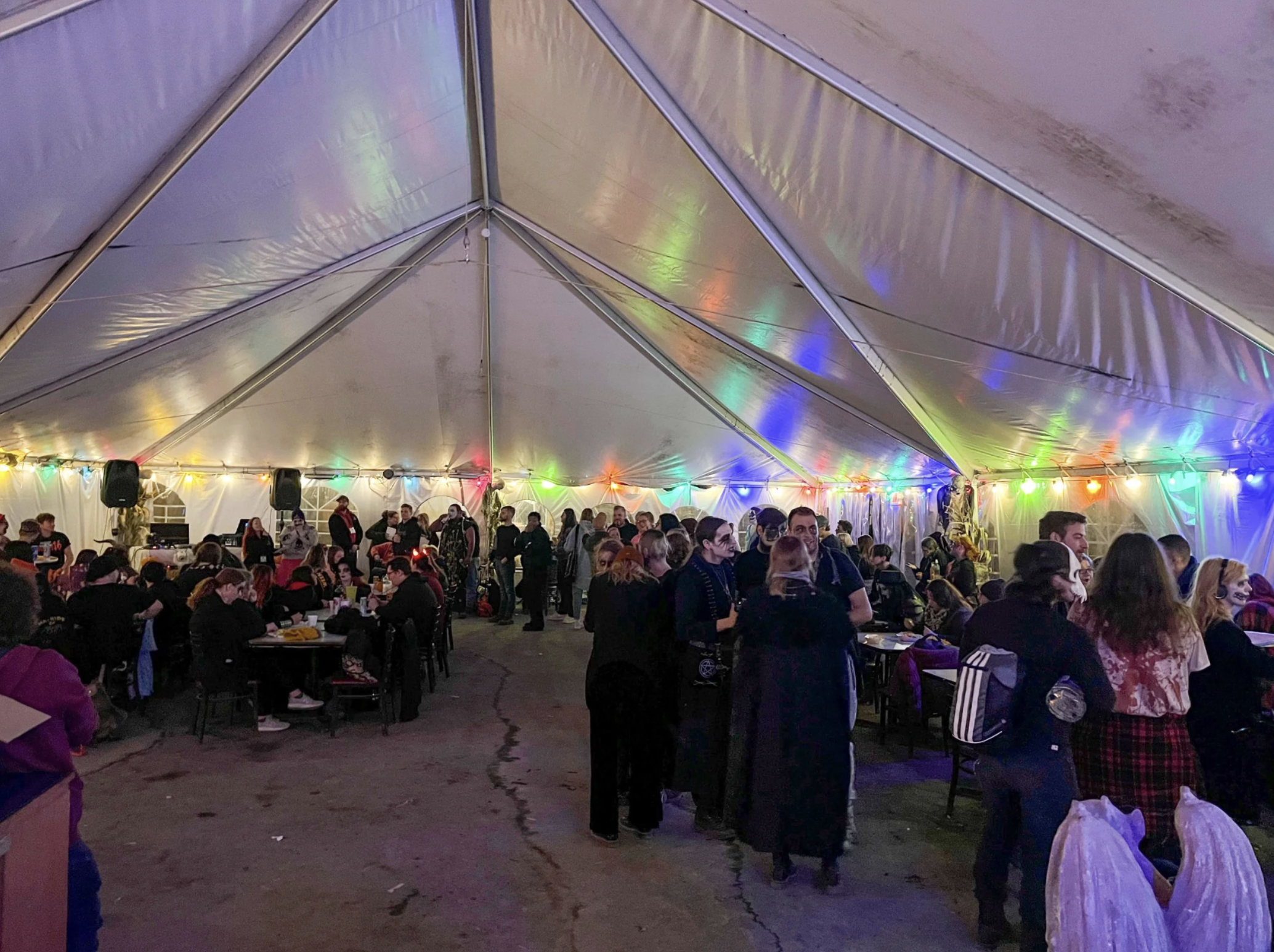 People gathered inside a large party tent decorated with colorful string lights, sitting at tables and standing.
