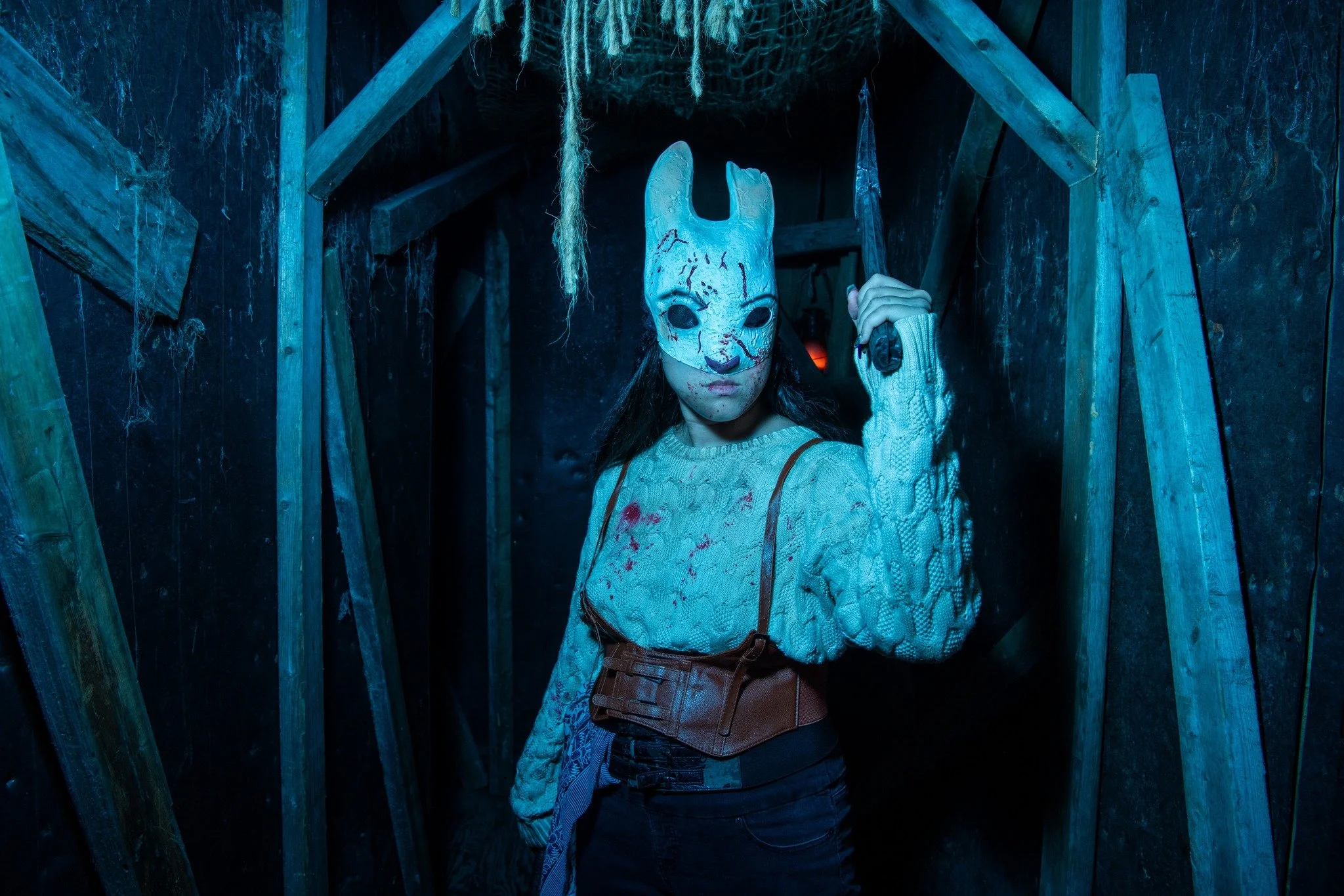 Person wearing a creepy rabbit mask and holding a weapon, standing inside a dark, wooden, haunted house-like structure.