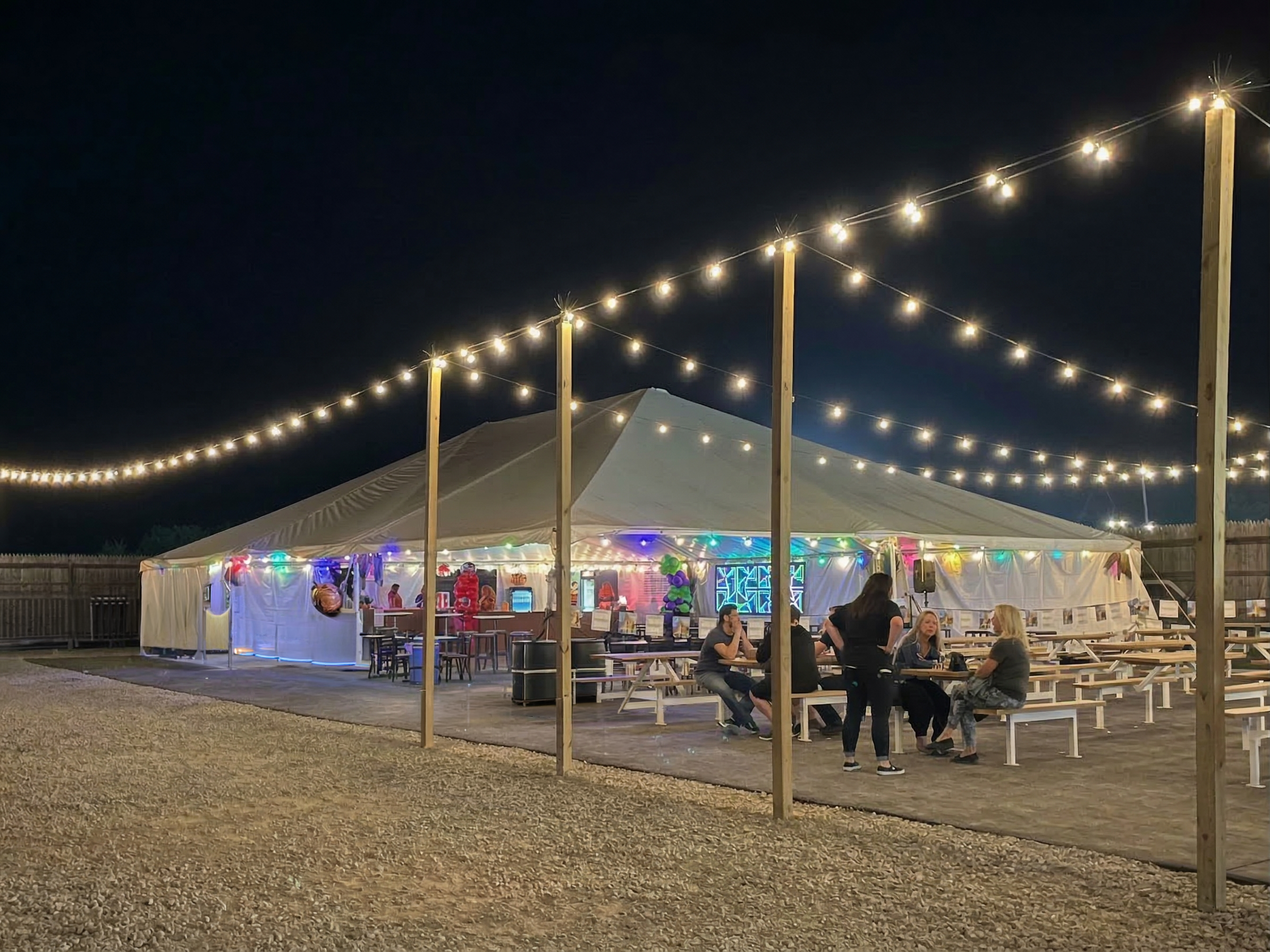 Night view of a large party tent decorated with string lights, colorful balloons, and LED lights. People are seated at picnic tables outside the tent, with some standing and talking. The ground is gravel in the foreground, and there is a wooden fence in the background.