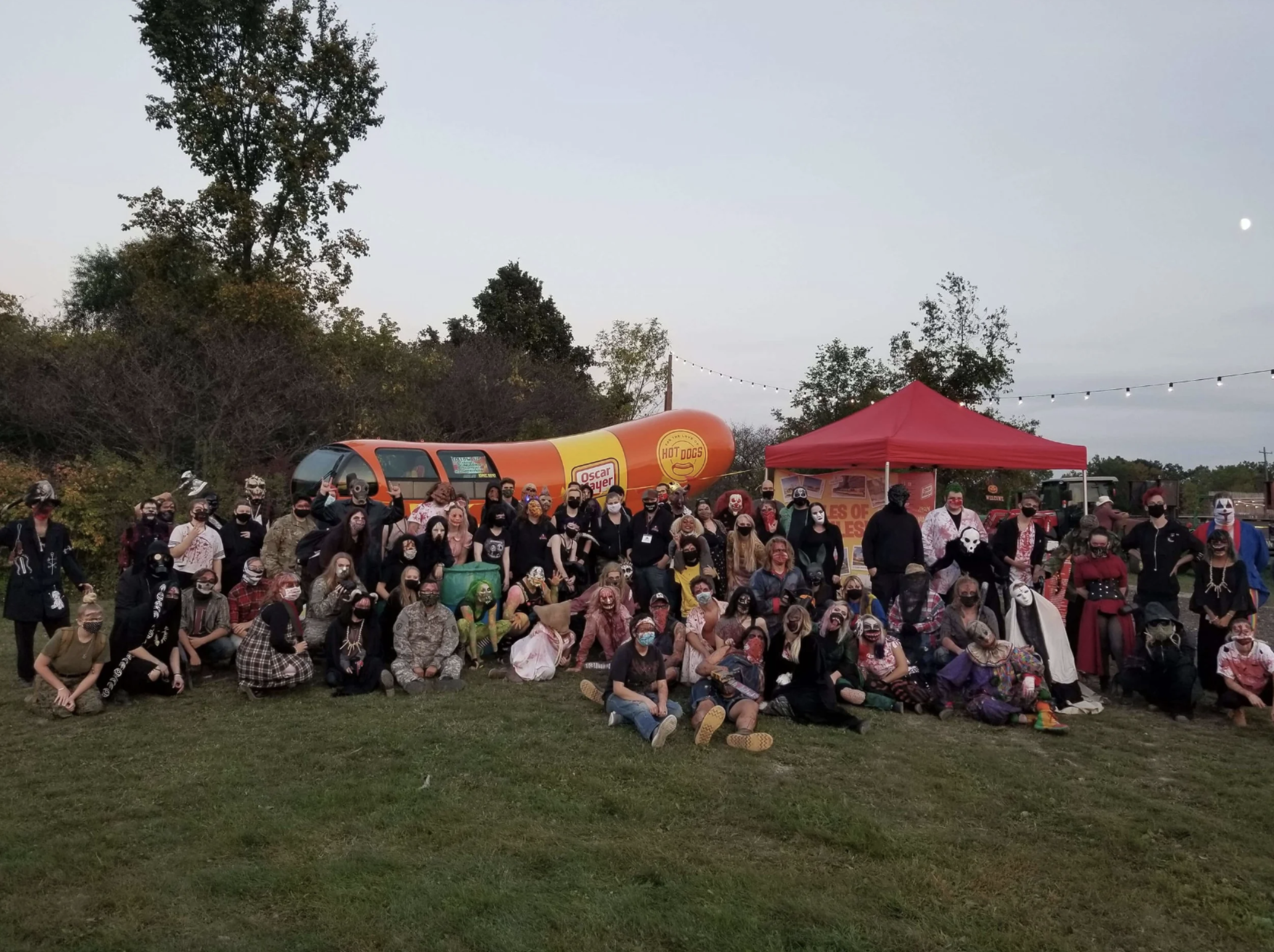 Large group of people dressed in Halloween costumes and masks, gathered outdoors at dusk, with a red tent, a large hot dog-shaped trailer, and string lights overhead.