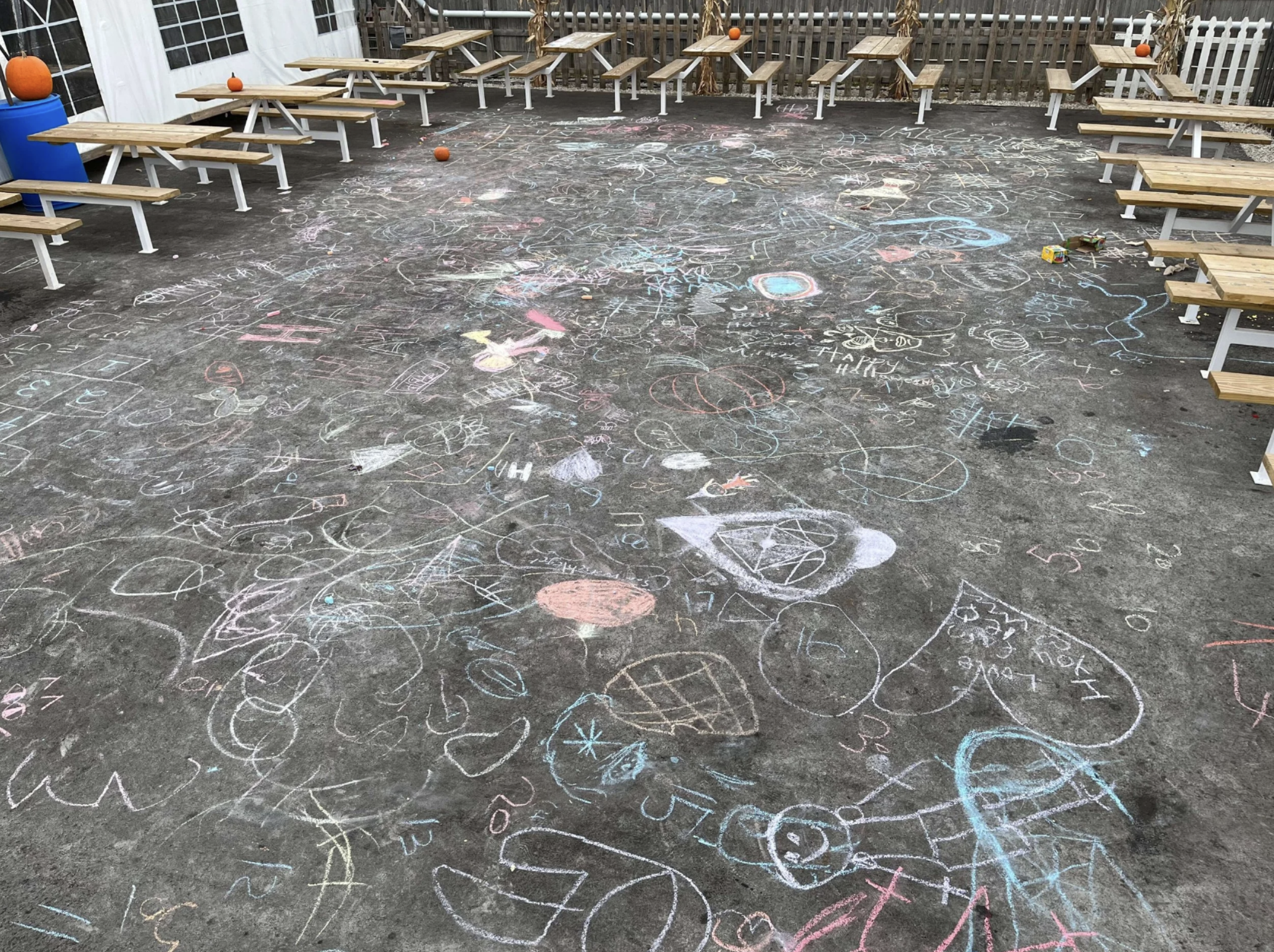 School playground with chalk drawings and scribbles on the ground, surrounded by picnic tables and a white fence.
