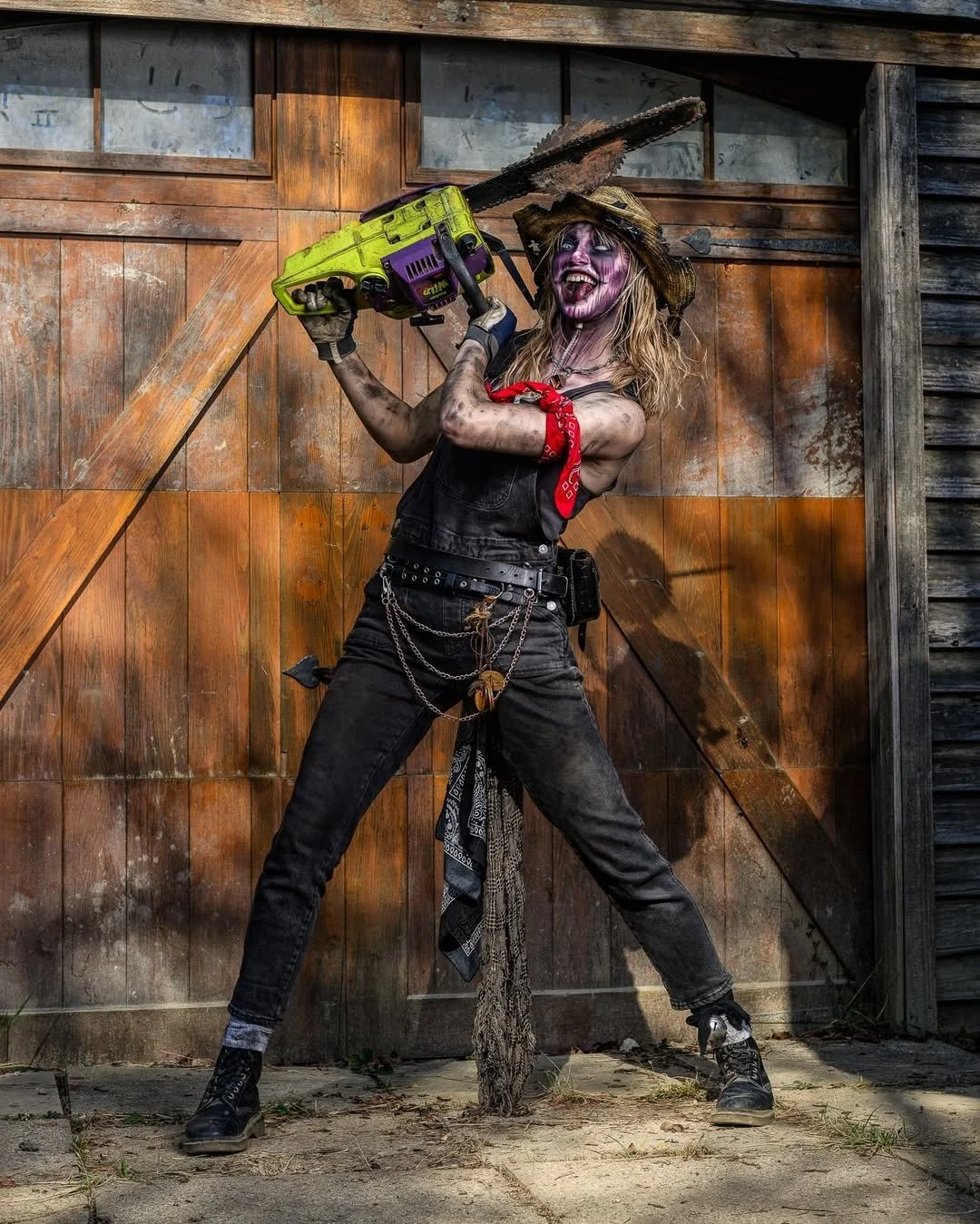 A woman dressed as a scary zombie given a sausage, holding a chainsaw, standing in front of a rustic wooden barn.