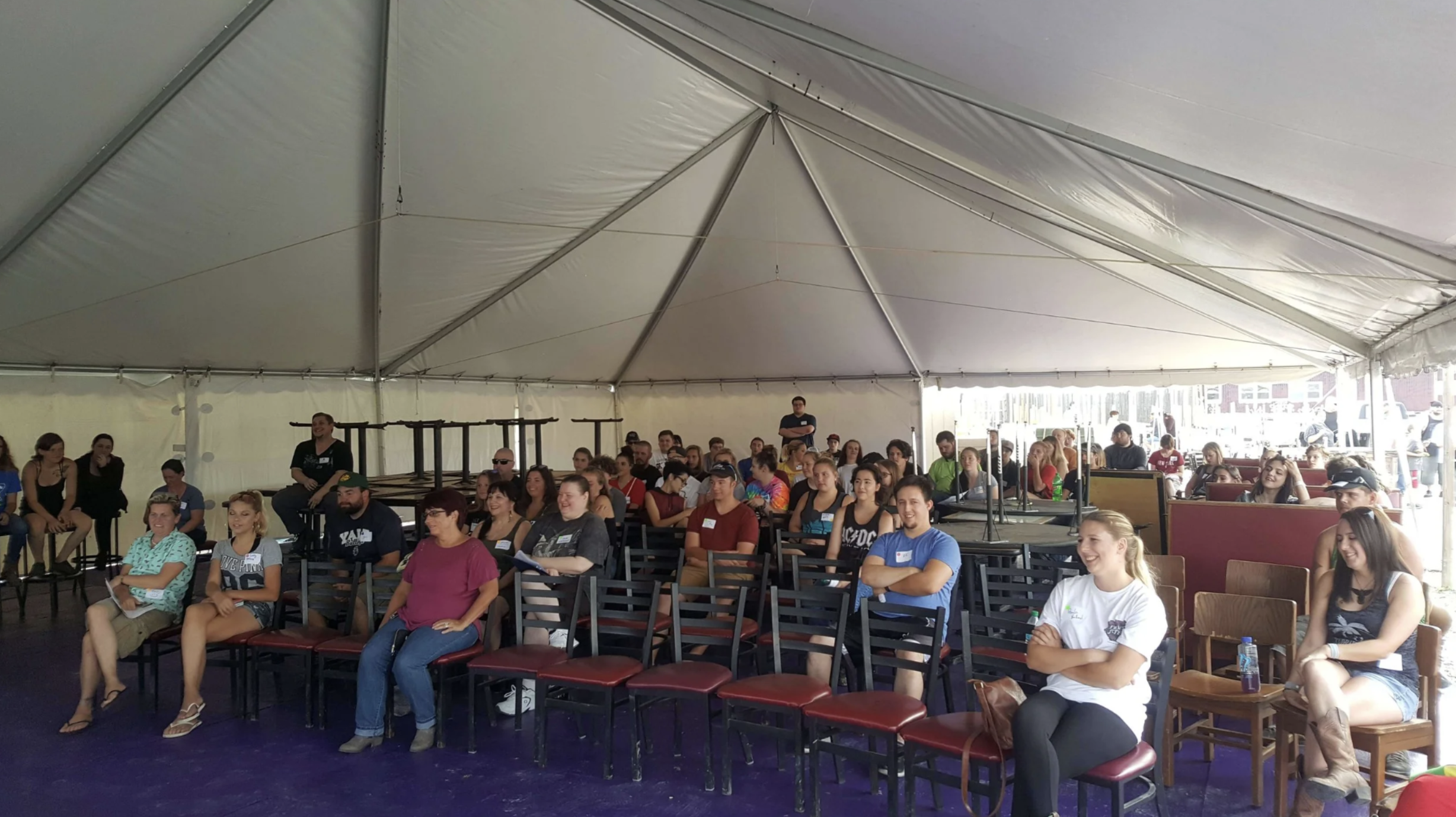 Group of people sitting and smiling inside a large white event tent with chairs and tables, some outdoors visible outside the tent.
