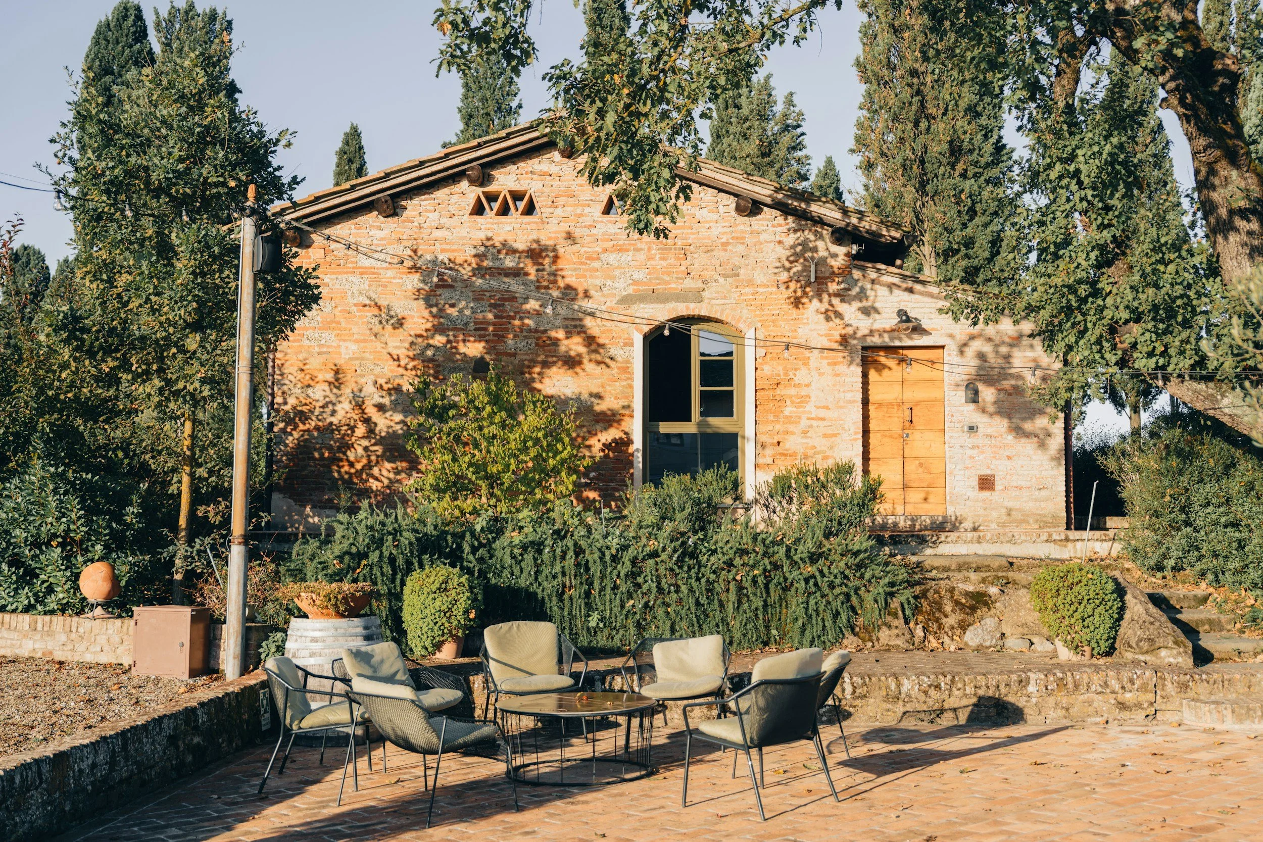 A brick house with a sloped roof, surrounded by green trees and bushes, featuring a patio with outdoor chairs and a table in the foreground.