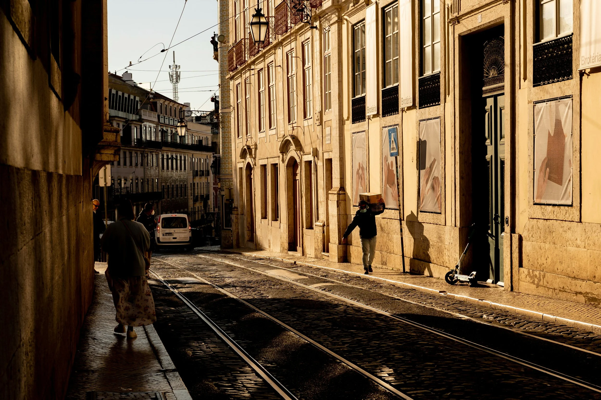 A narrow cobblestone street in a European city with tram tracks. Pedestrians walk along the sidewalk, one carries a box on their shoulder, and a scooter is parked near a building. The buildings have large windows and ornate details, and wires crisscross overhead. The scene is bathed in warm sunlight.