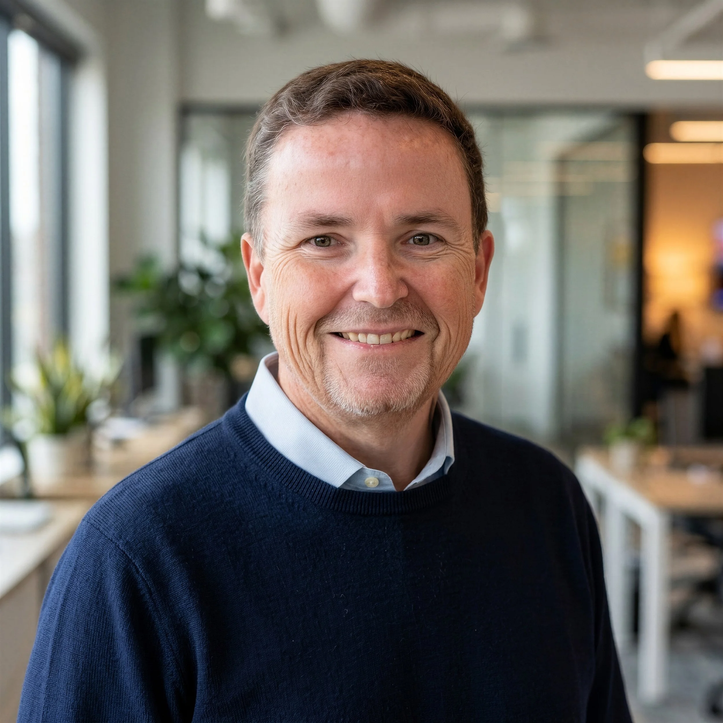Smiling middle-aged man in a navy sweater and collared shirt posing in a modern office