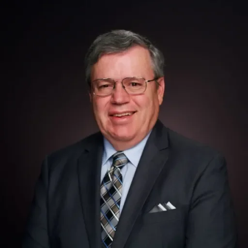 A middle-aged man with gray hair and glasses, dressed in a dark suit and a plaid tie, smiling against a dark background.