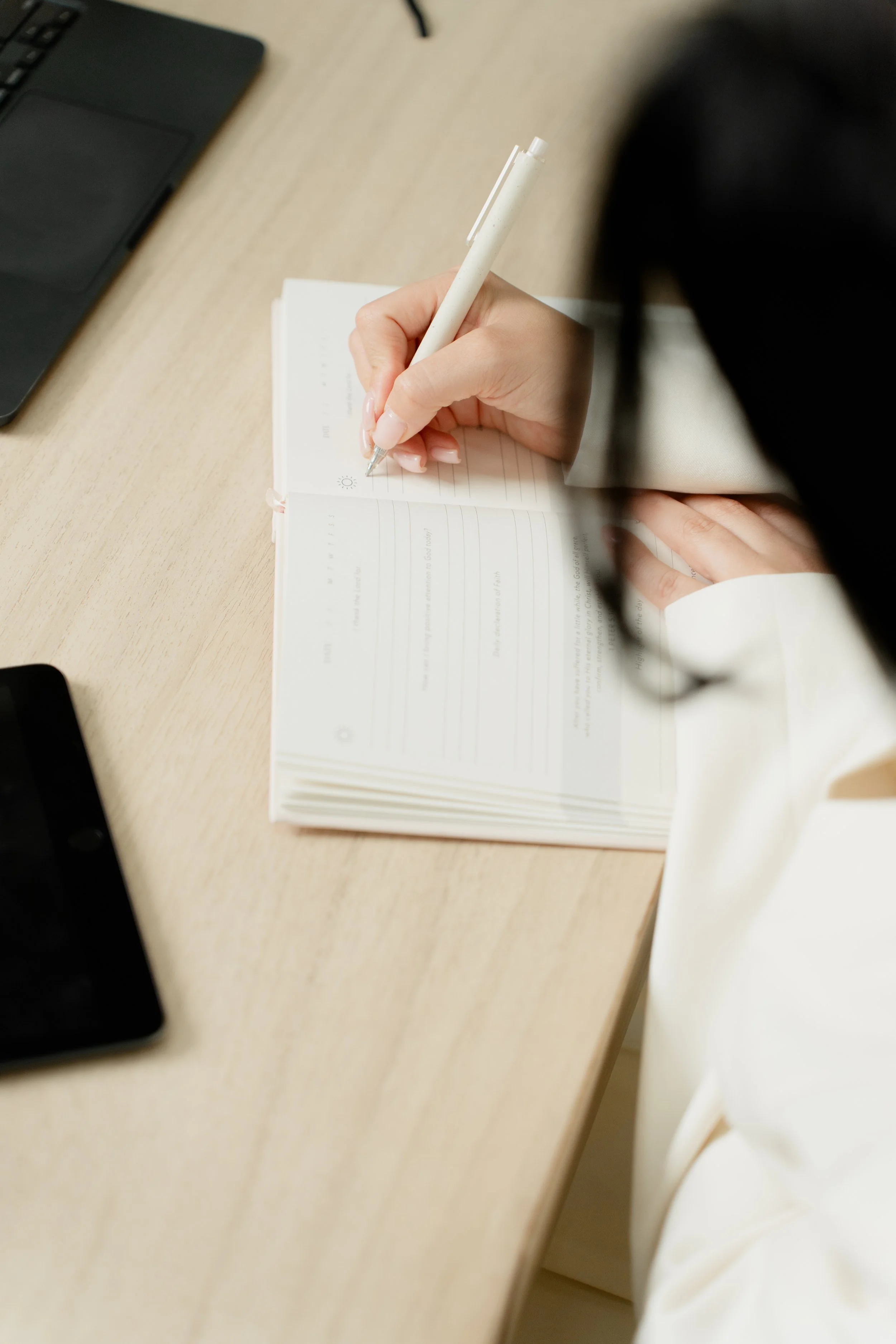 Person writing in a notebook with a pen on a wooden desk, with smartphones and a laptop nearby.