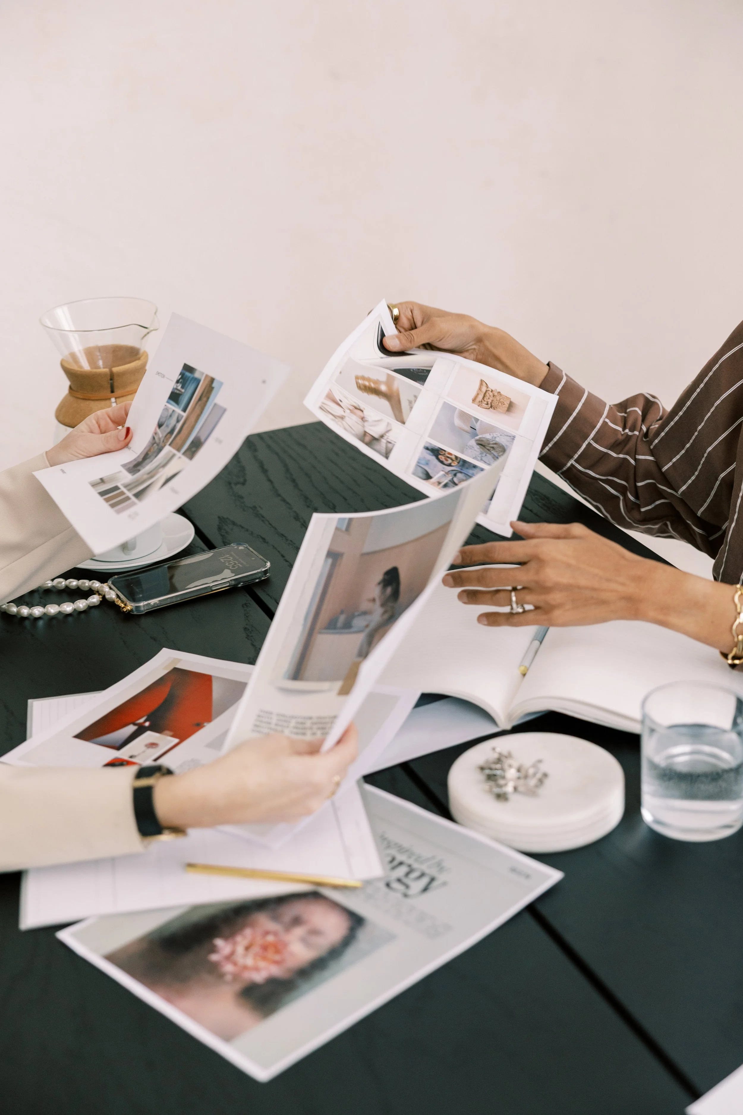 Three people sitting at a table looking at printed photographs and magazines, with a glass of water and a bowl on the table, and one person holding a printed photograph.