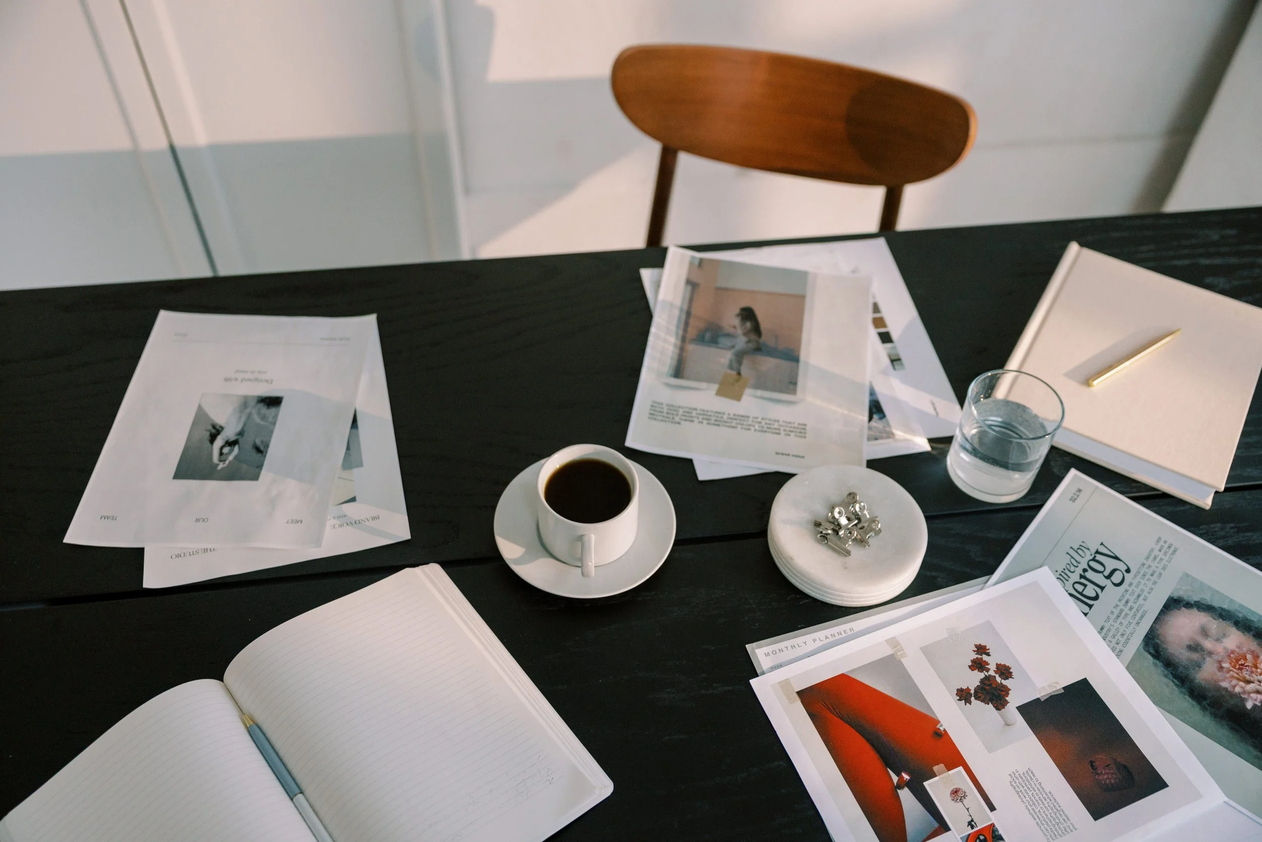 A black table with various papers, a notebook, a pen, a cup of coffee, a glass of water, a small bowl of clips, a closed white notebook, and a magazine, with a wooden chair in the background.