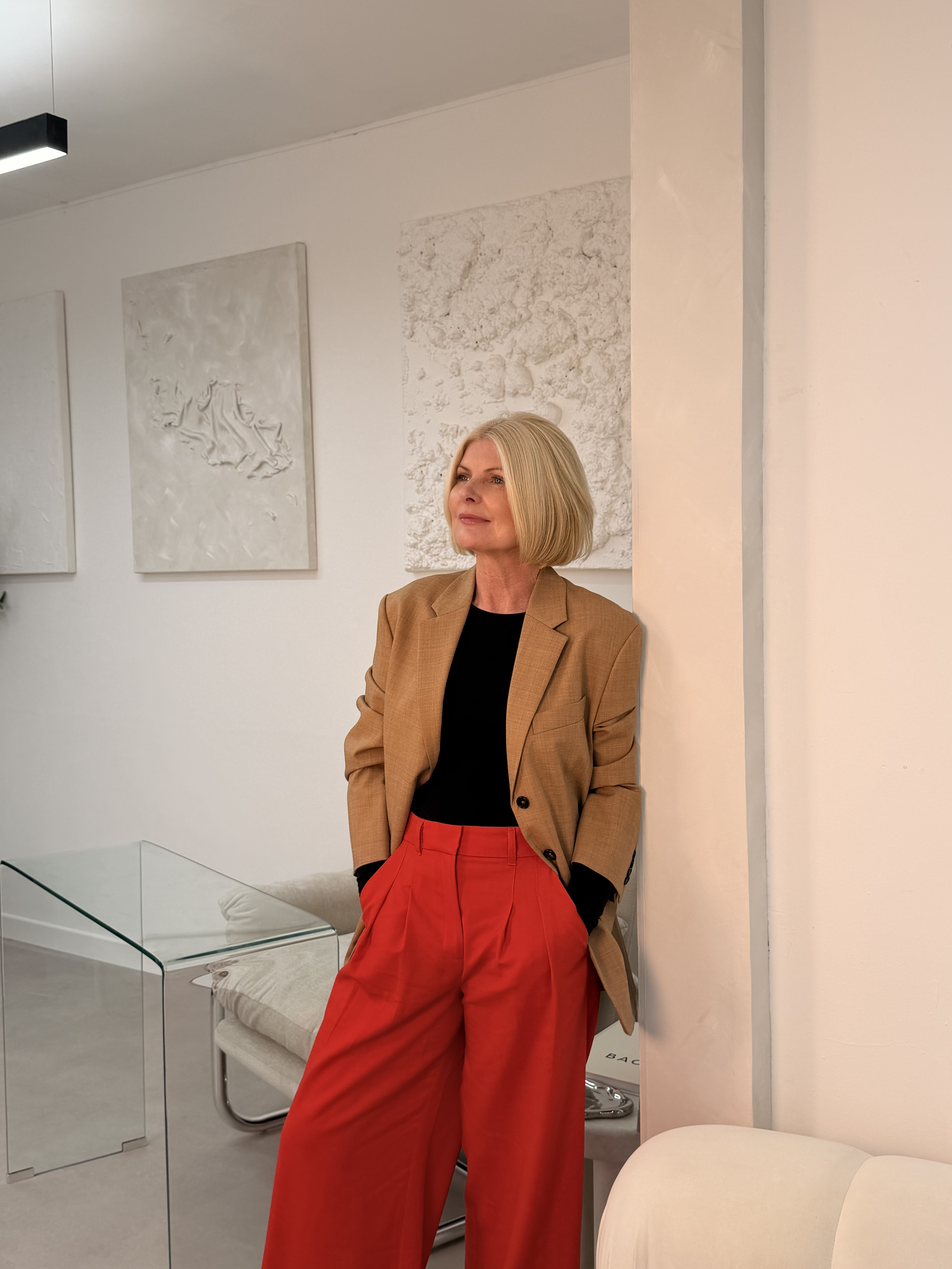 Alex Darby with shoulder-length blonde hair wearing a tan blazer, black top, and red wide-leg trousers, standing in a modern, white art gallery or interior space leaning against a wall.