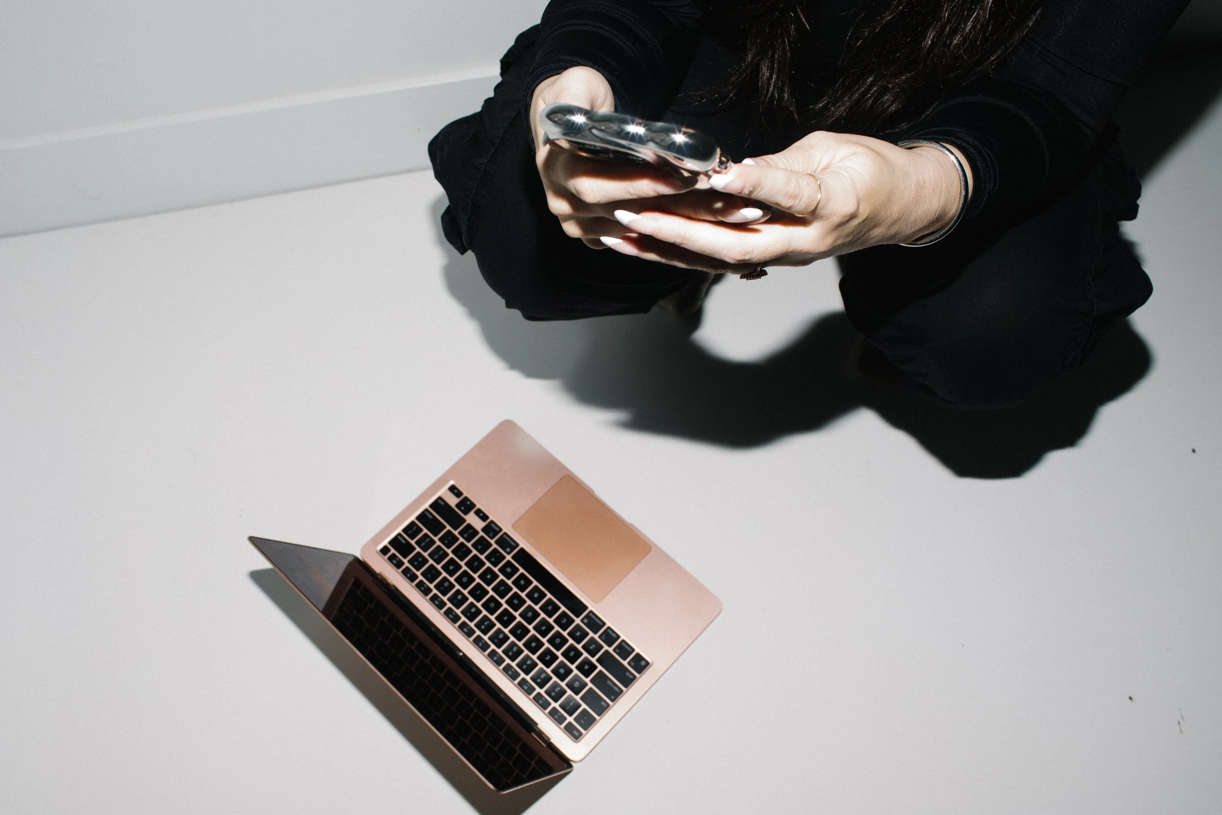 A person with long dark hair is sitting at a white table, looking at a smartphone with both hands. A small, rose gold laptop is open on the table. The person is wearing a black long-sleeve shirt or jacket, and has a bracelet and a ring on their left hand.