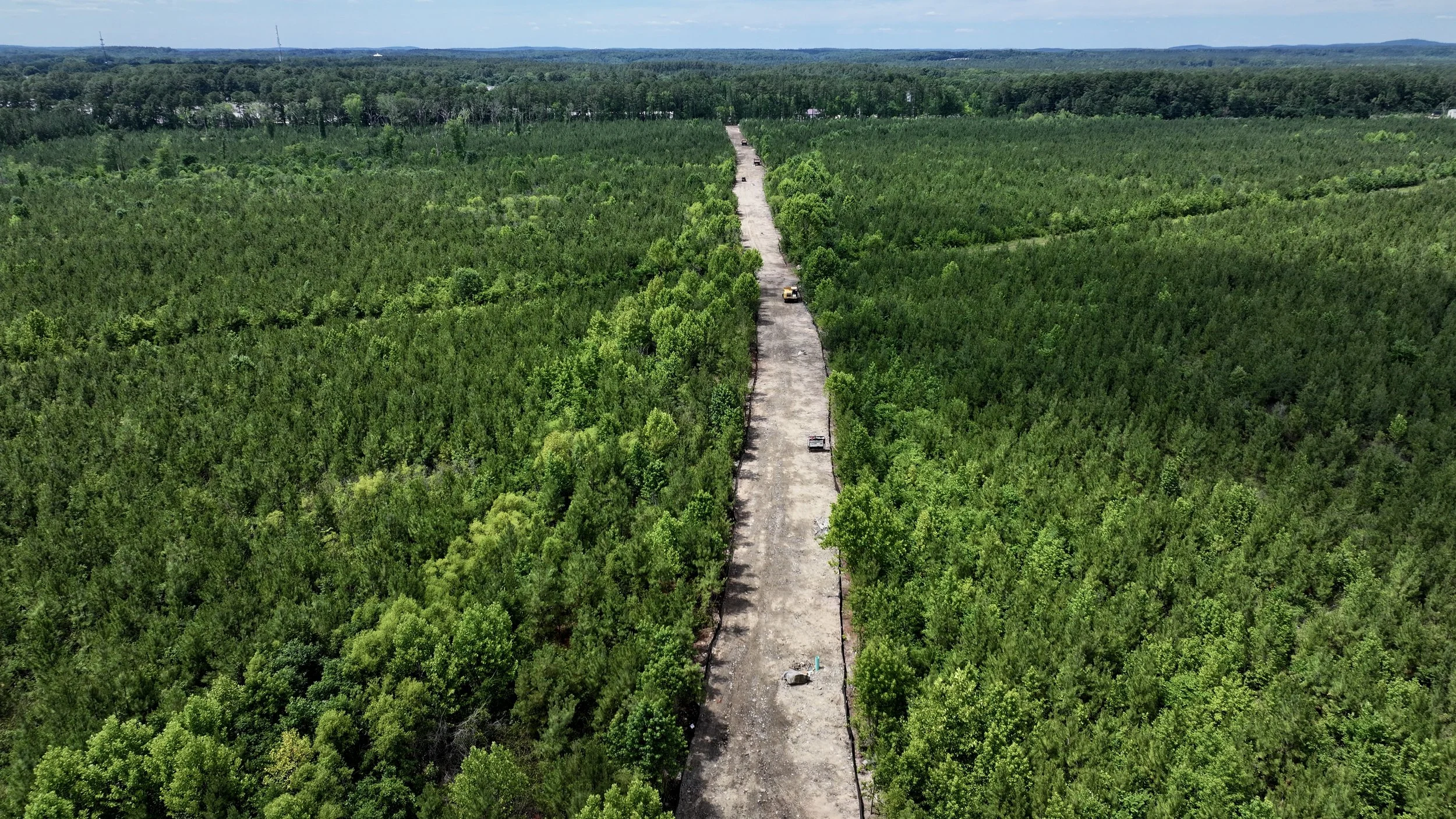 Aerial view of a narrow construction or dirt road running through a dense, green forest under a partly cloudy sky.