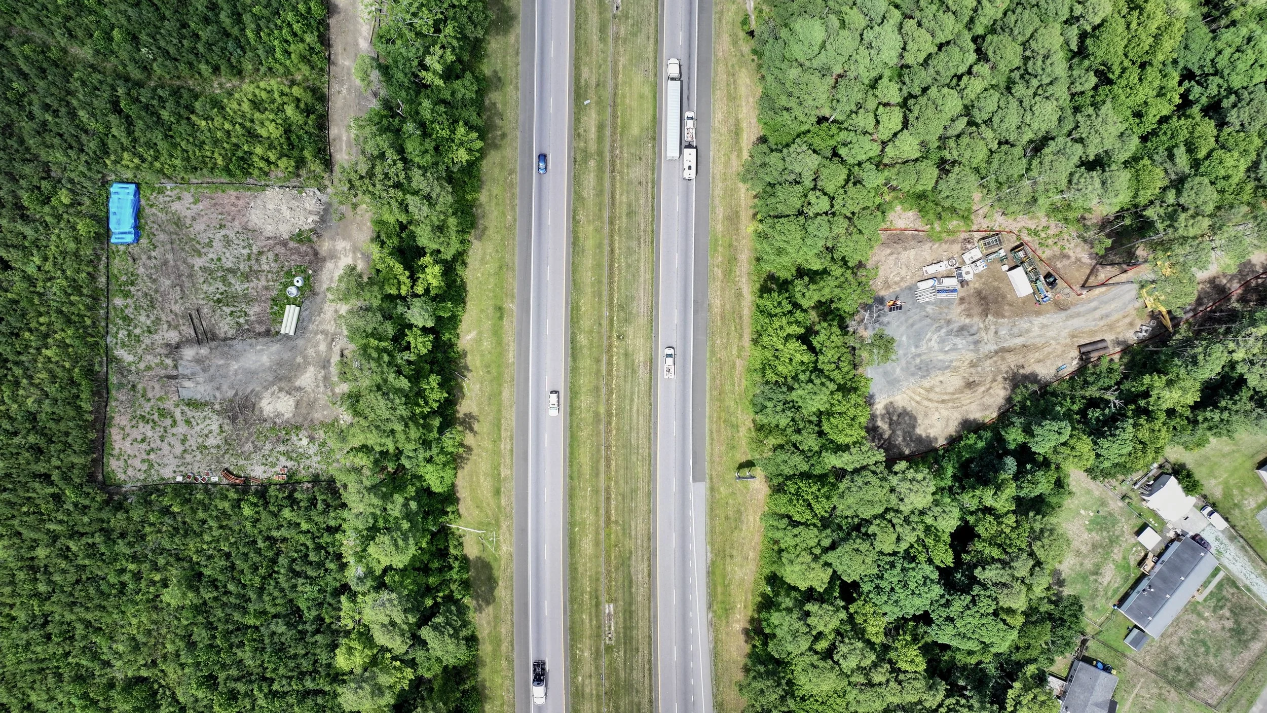An aerial view of a highway crossing through a forested area with two lanes of traffic traveling in each direction. Surrounding the highway are green trees, cleared lots, and some buildings.