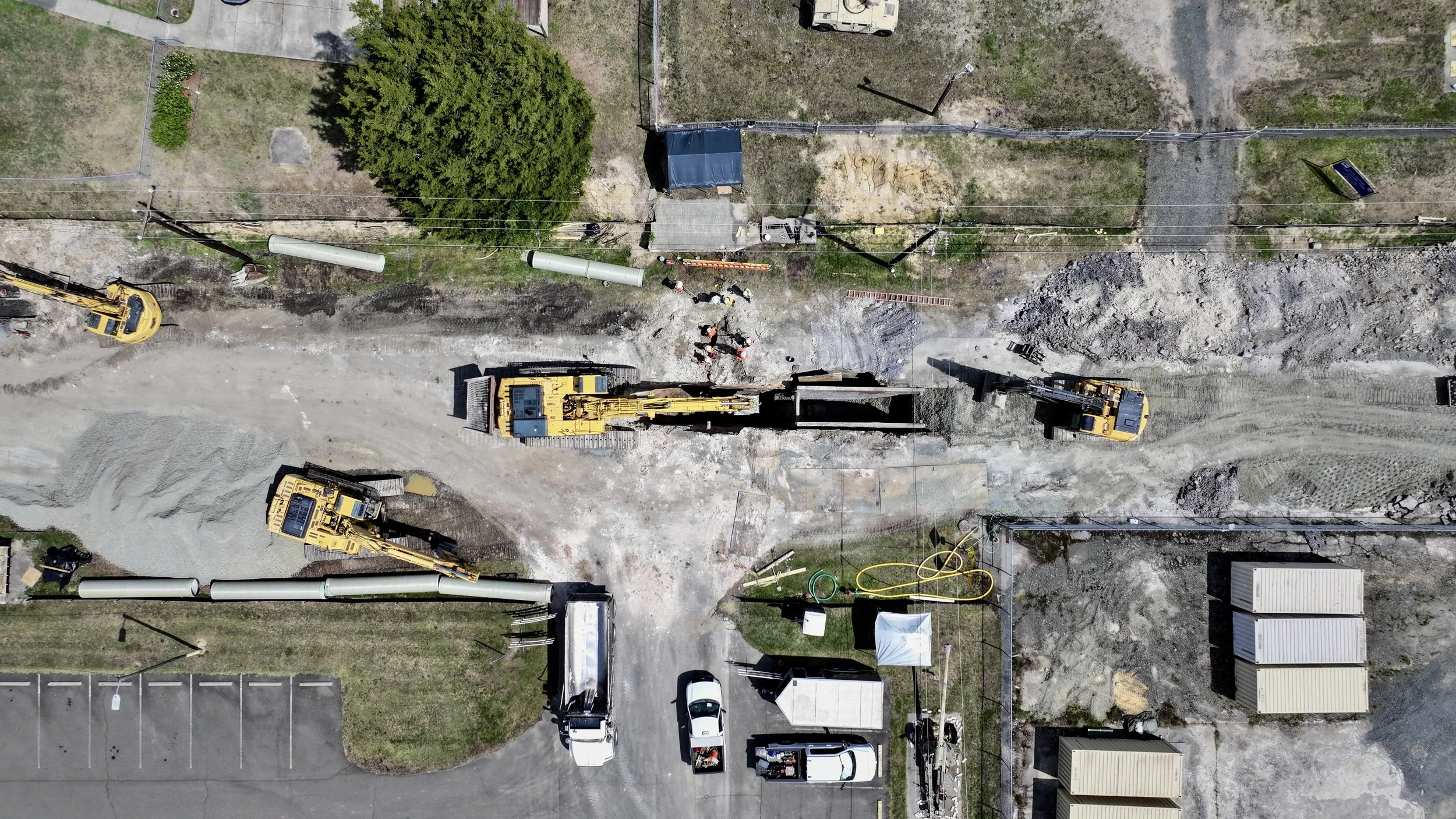 An aerial view of a construction site with excavators, construction workers, pipes, and machinery, with a fenced yard and parking area nearby.