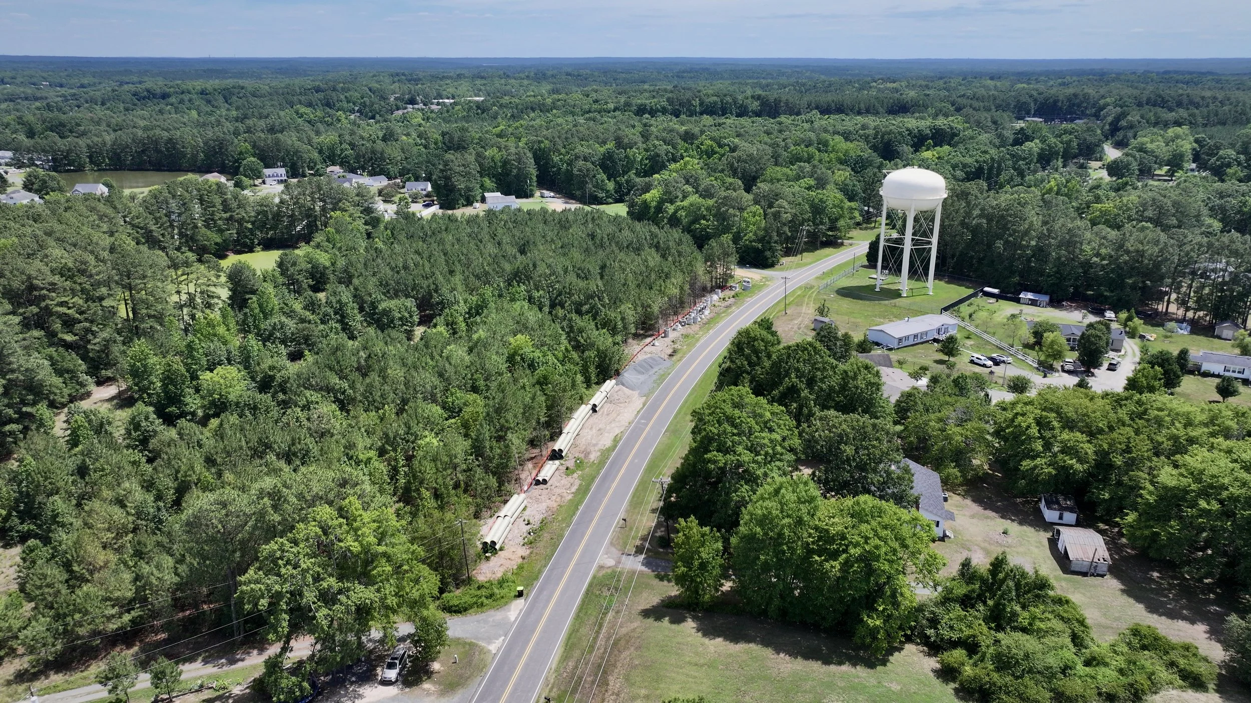 An aerial view of a rural area with a water tower, houses, trees, a winding road, and pipeline construction.