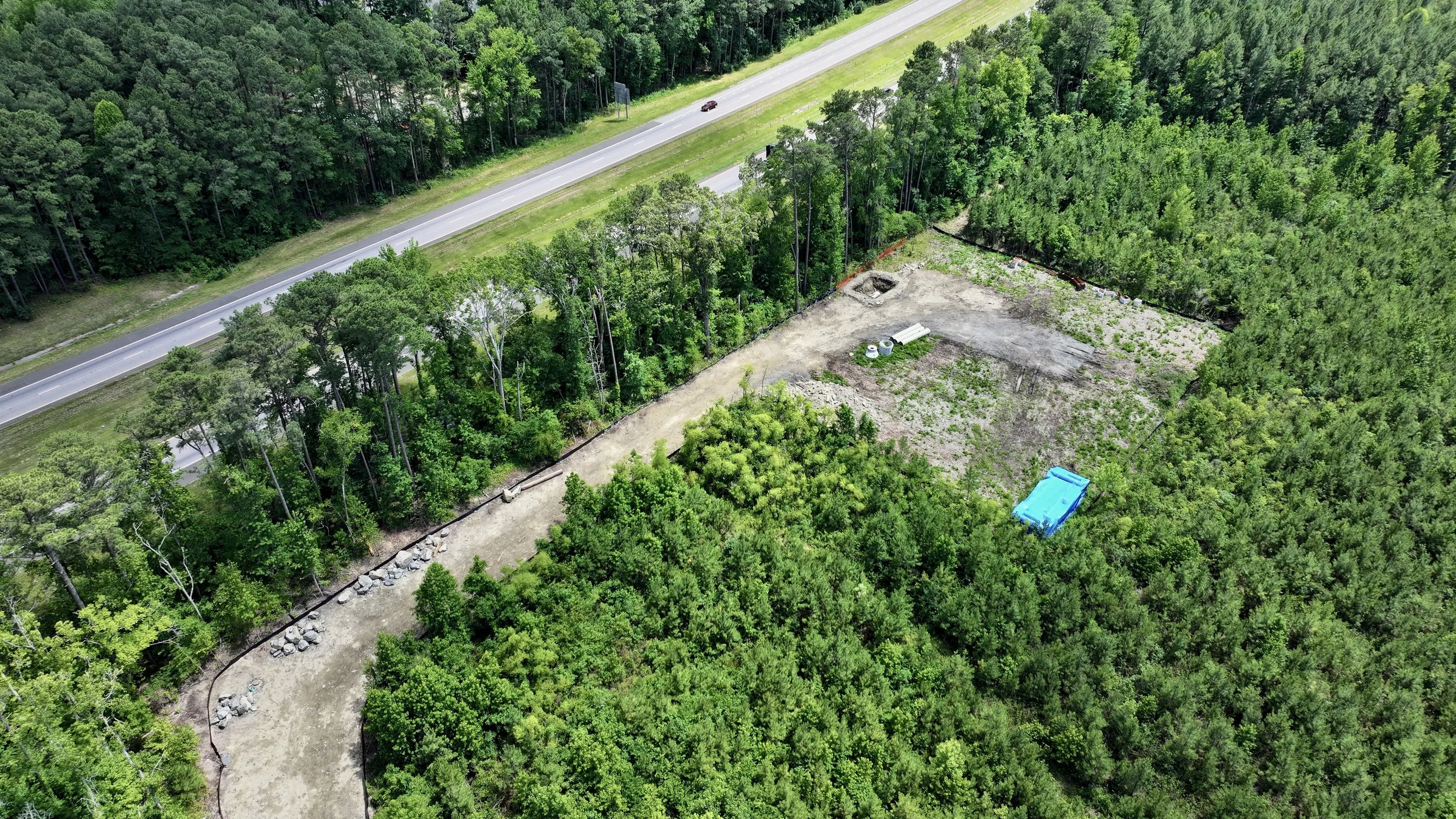 An aerial view of a construction site next to a two-lane road surrounded by dense green forest. The construction area has some dirt, rocks, a small trench, and construction materials, with a blue container or equipment visible in the lower right corn