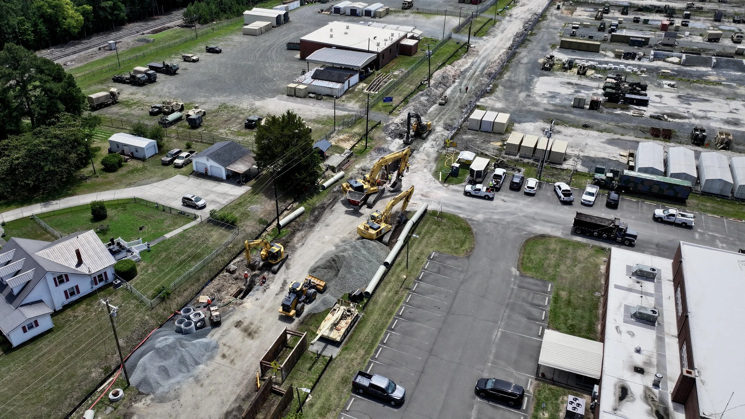 Construction site with excavators, construction workers, and ongoing road work; parked cars and surrounding buildings.
