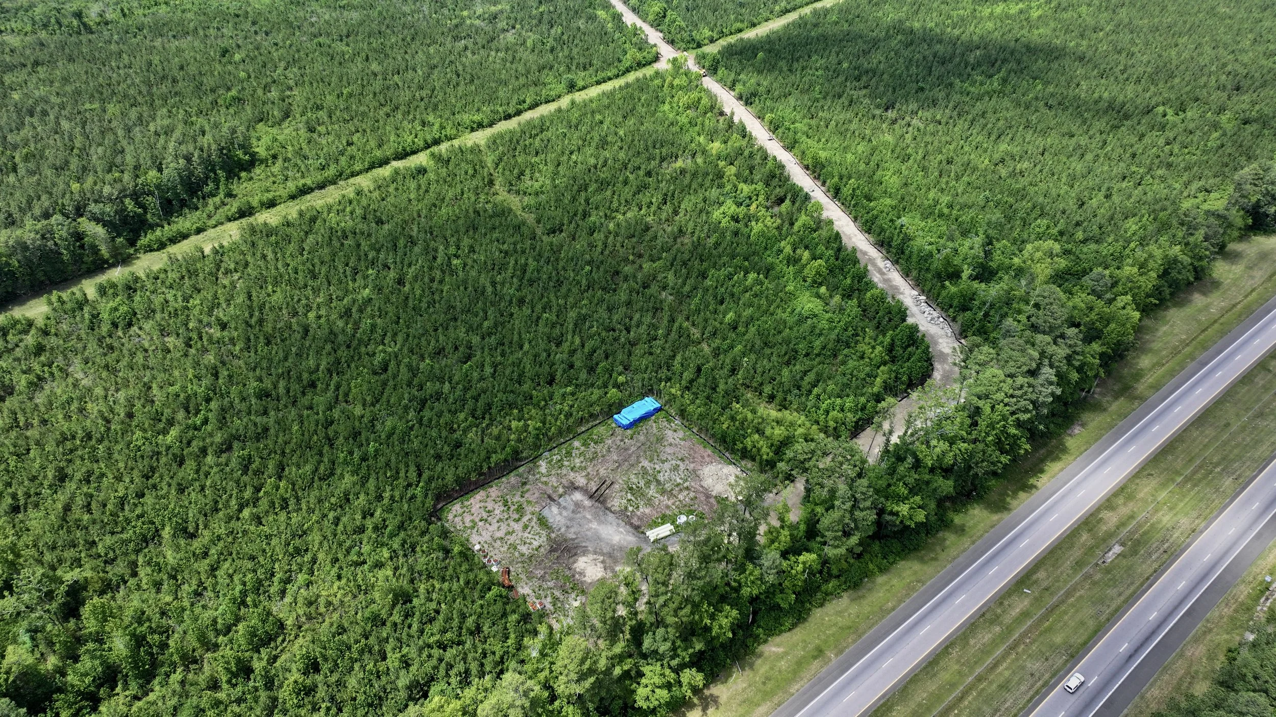Aerial view of a lush green forest with a dirt path running through it, alongside a paved road. There is a small blue structure and some equipment in a cleared area near the forest.