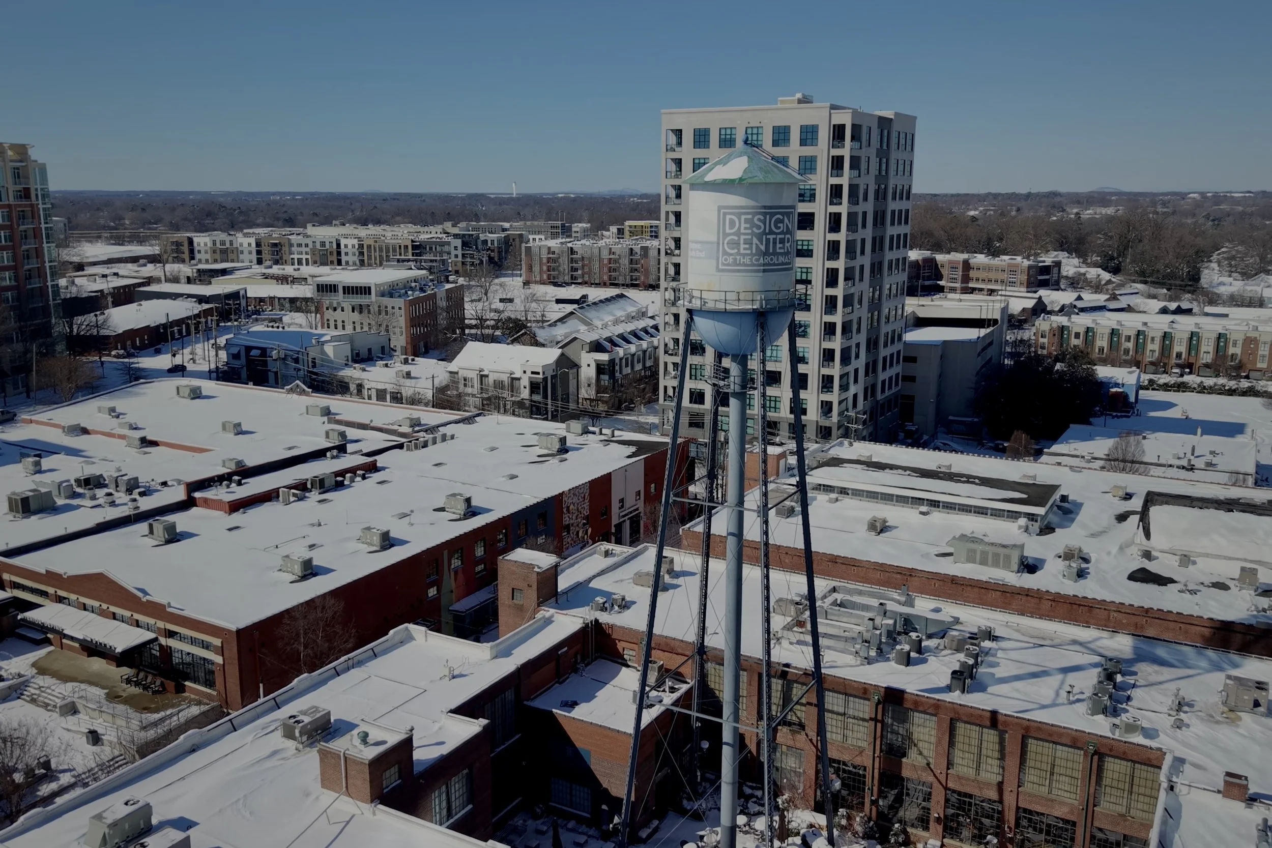 A cityscape during winter with snow-covered rooftops, a water tower labeled.'Design Center of the Carolinas,' and modern buildings under a clear blue sky.