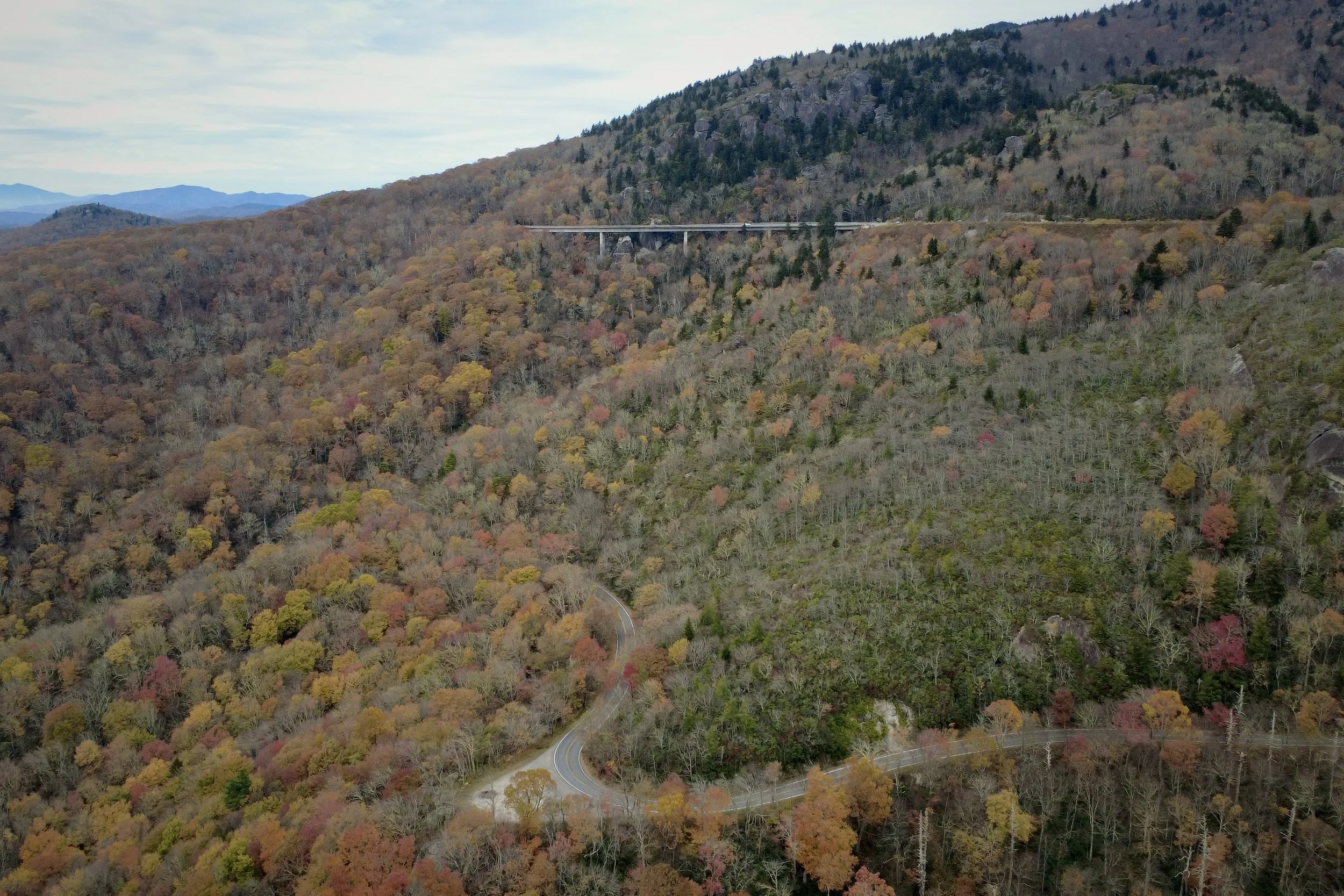 Aerial view of a mountainous landscape with a winding road and a bridge, covered with autumn-colored trees.