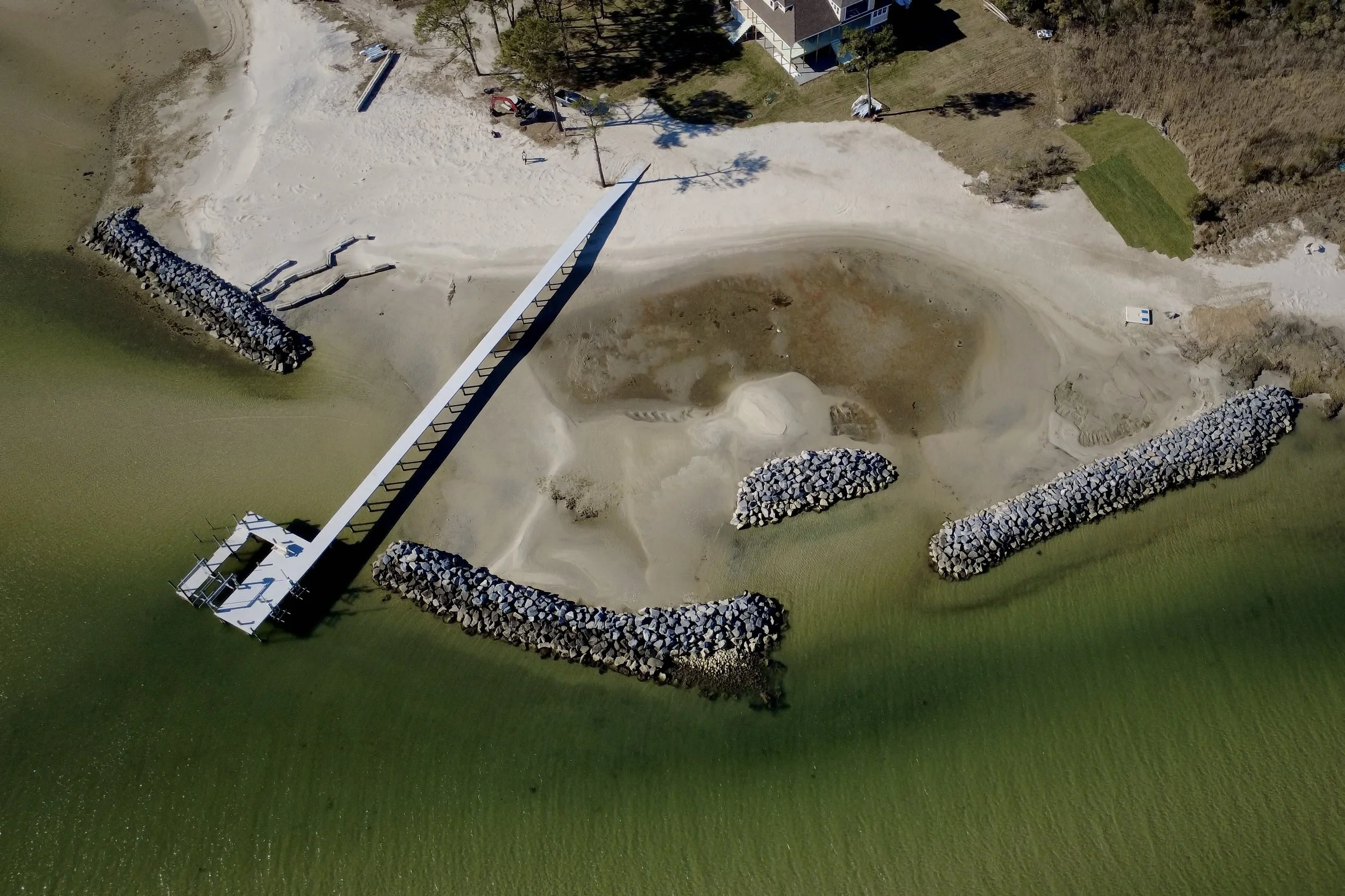 Aerial view of a beach area with a long pier extending into the water, surrounded by rock breakwaters and sandy shores with some grass and trees nearby.