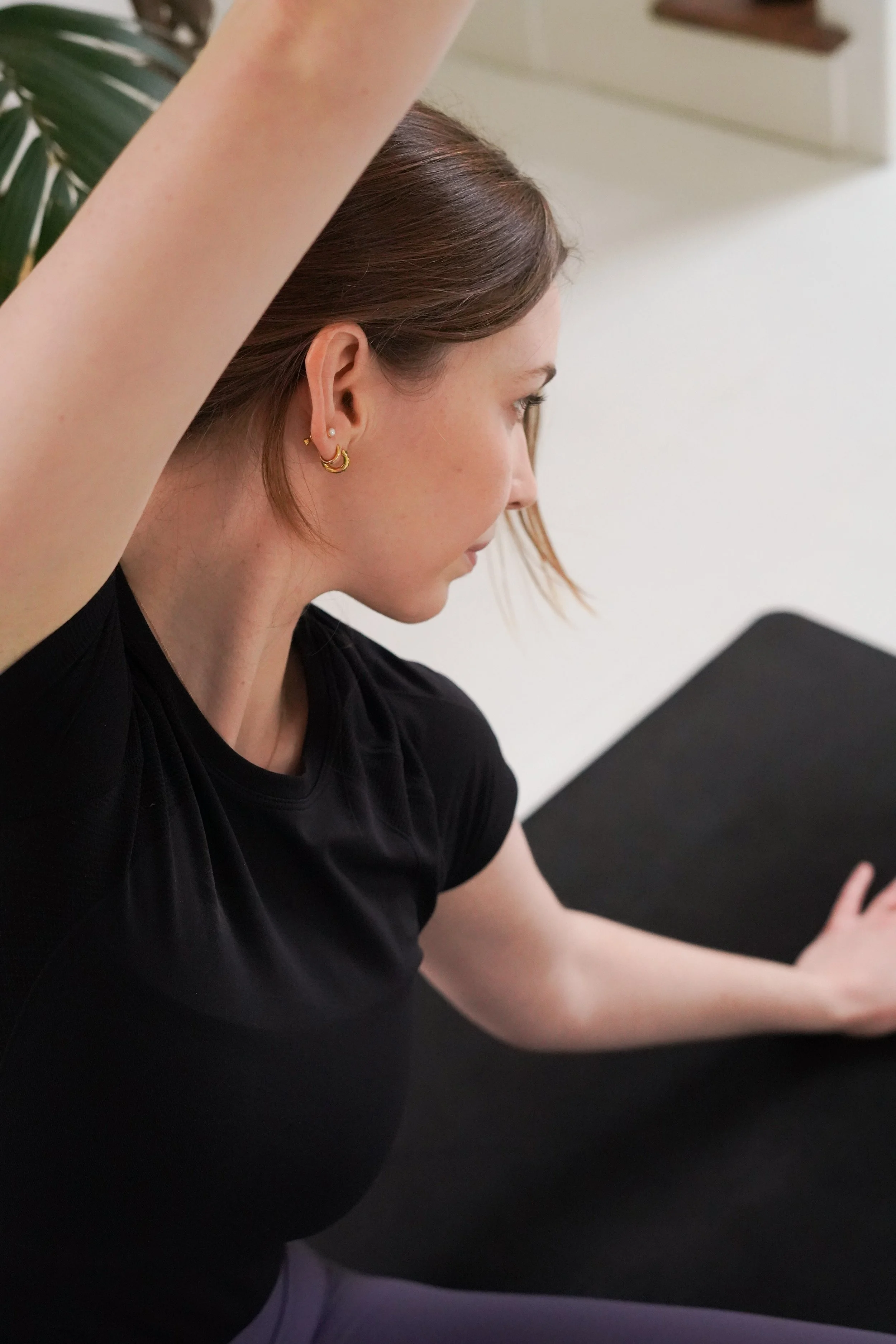 Woman practicing yoga or stretching on a black mat indoors, with her arm raised and head turned to the side.