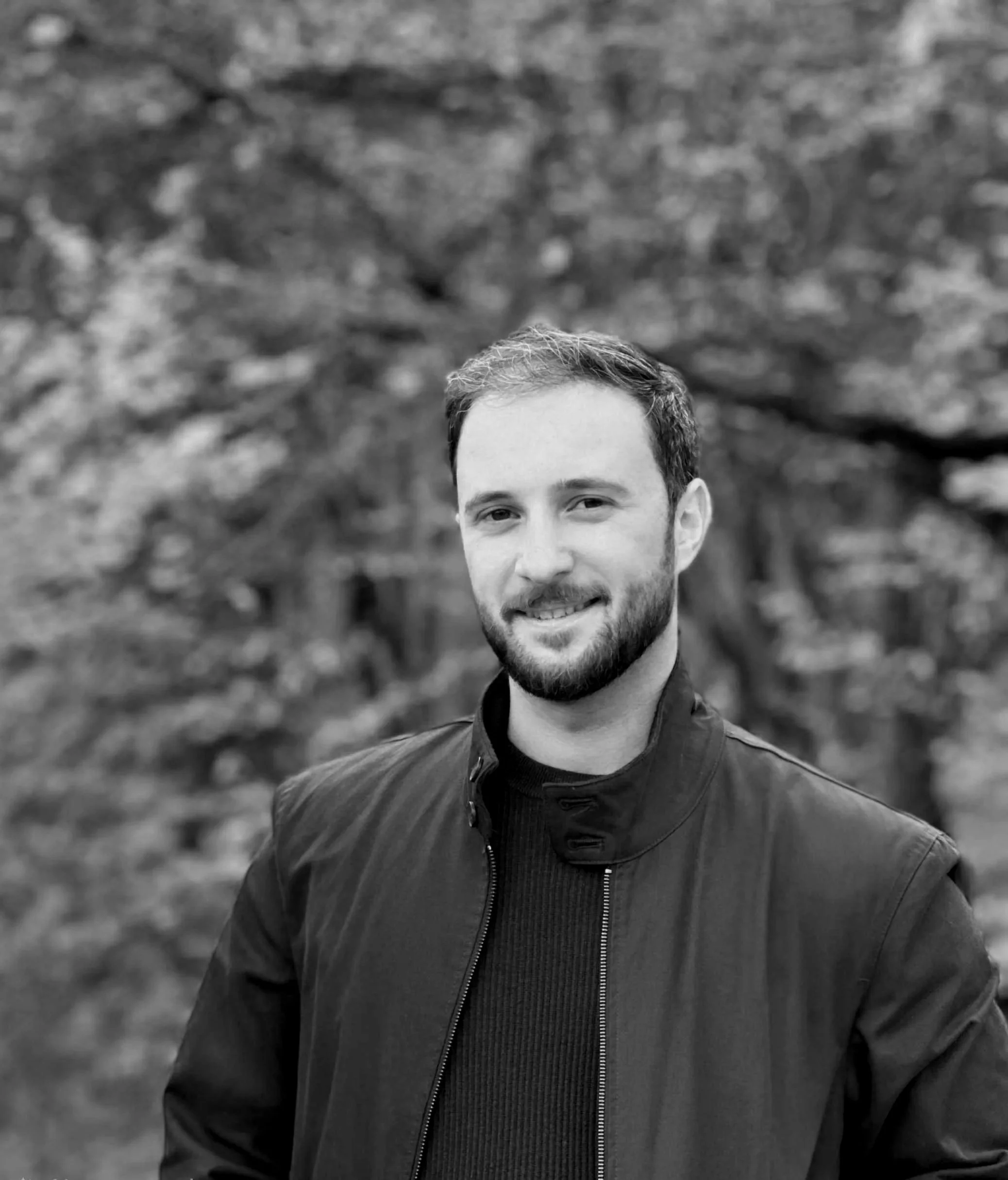 A black and white portrait of a man with short hair and a beard, smiling, wearing a dark jacket, outdoors with blurred trees in the background.