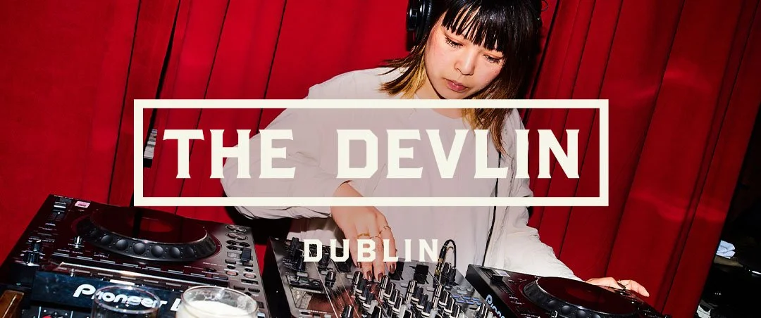 A young woman DJ wearing headphones mixes music at a DJ booth with Pioneer equipment, in front of a red wall, with a glass of beer in the foreground.