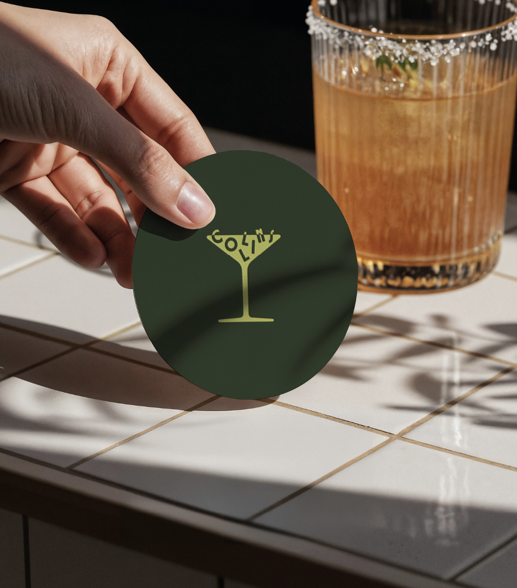 A person's hand holding a green circular sticker with a yellow martini glass design made of the word 'Cocktail'. In the background, there is a glass of brown mixed drink with ice, garnished with a lime wedge, sitting on a tiled surface.