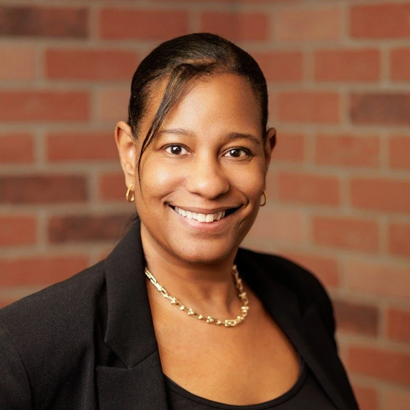 Professional woman wearing a black blazer and gold jewelry, smiling in front of a brick wall.