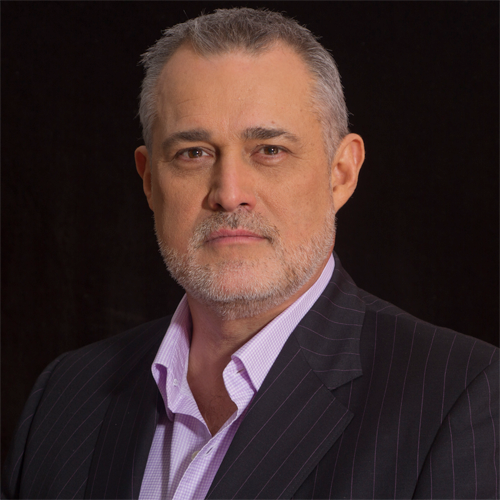 Portrait of a middle-aged man with gray hair and beard wearing a dark pinstripe suit and light purple collared shirt against a dark background.