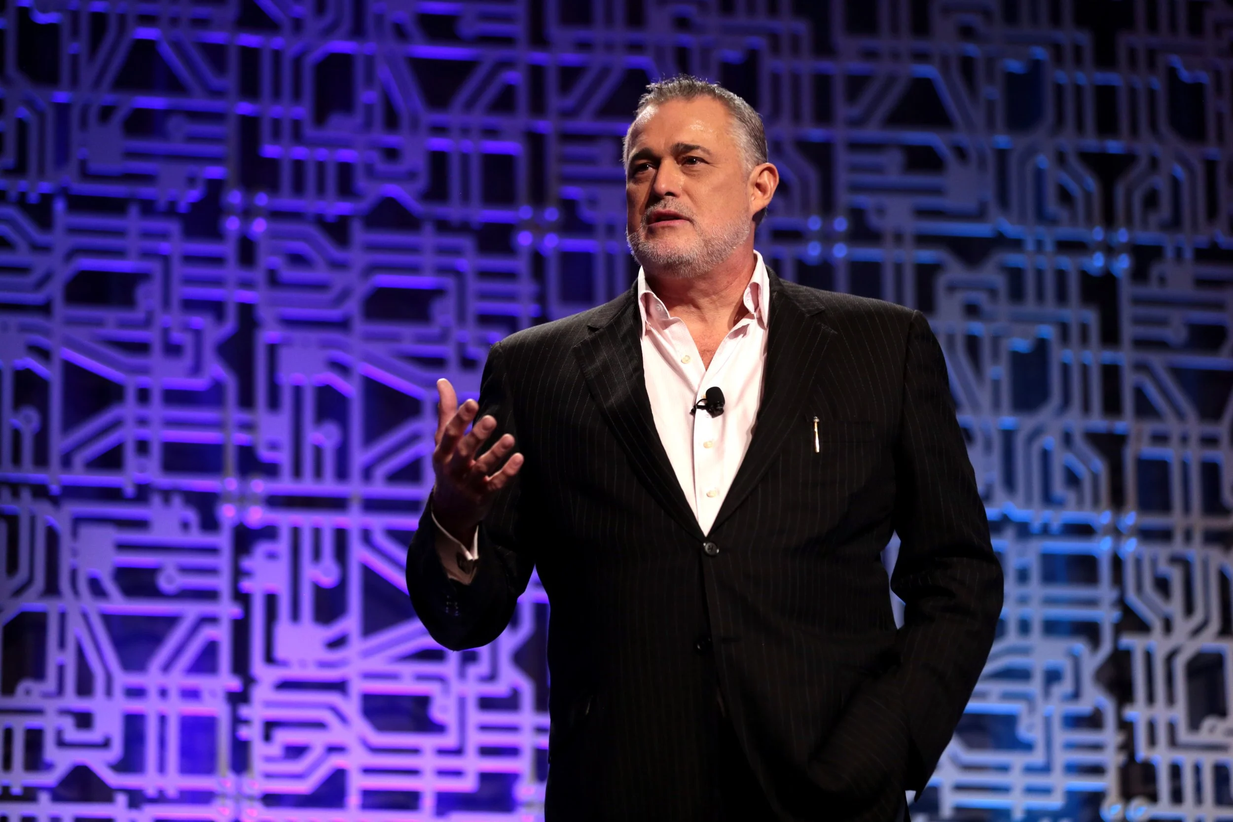 A man in a black pinstripe suit and white shirt speaking or presenting on stage, with a blue and purple geometric patterned background.