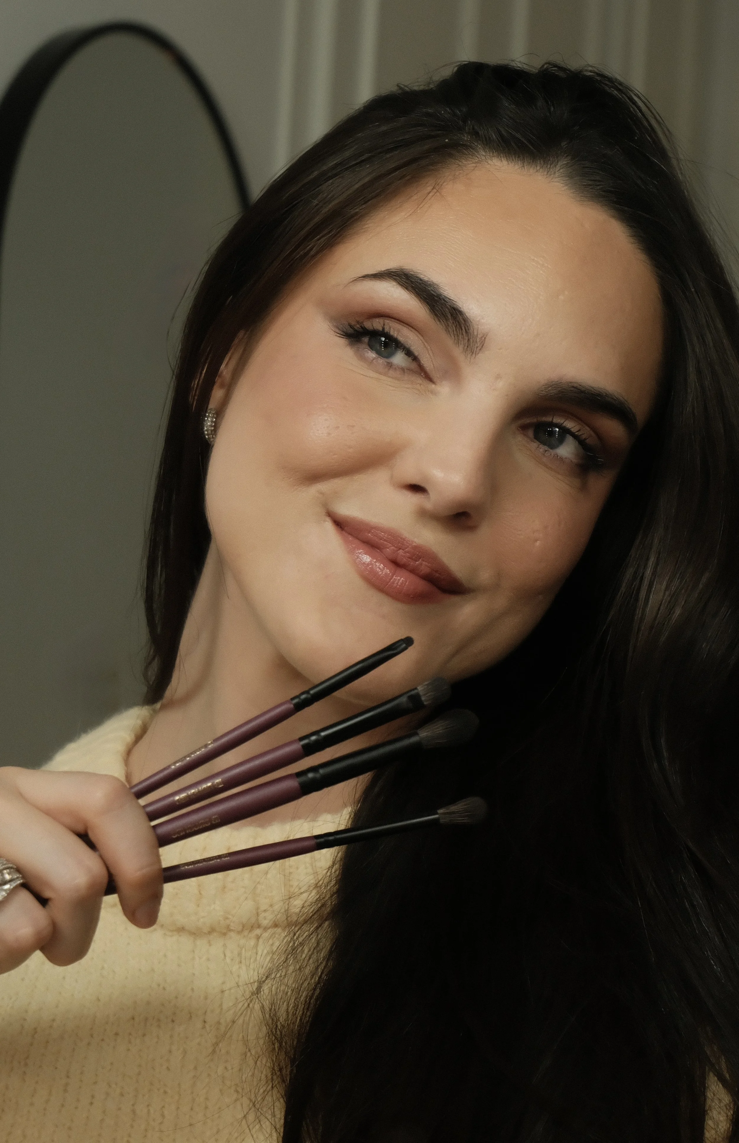 Mujer con cabello oscuro, con maquillaje y labios de color marrón, sosteniendo varios pinceles de maquillaje en su mano, sonriendo ligeramente.