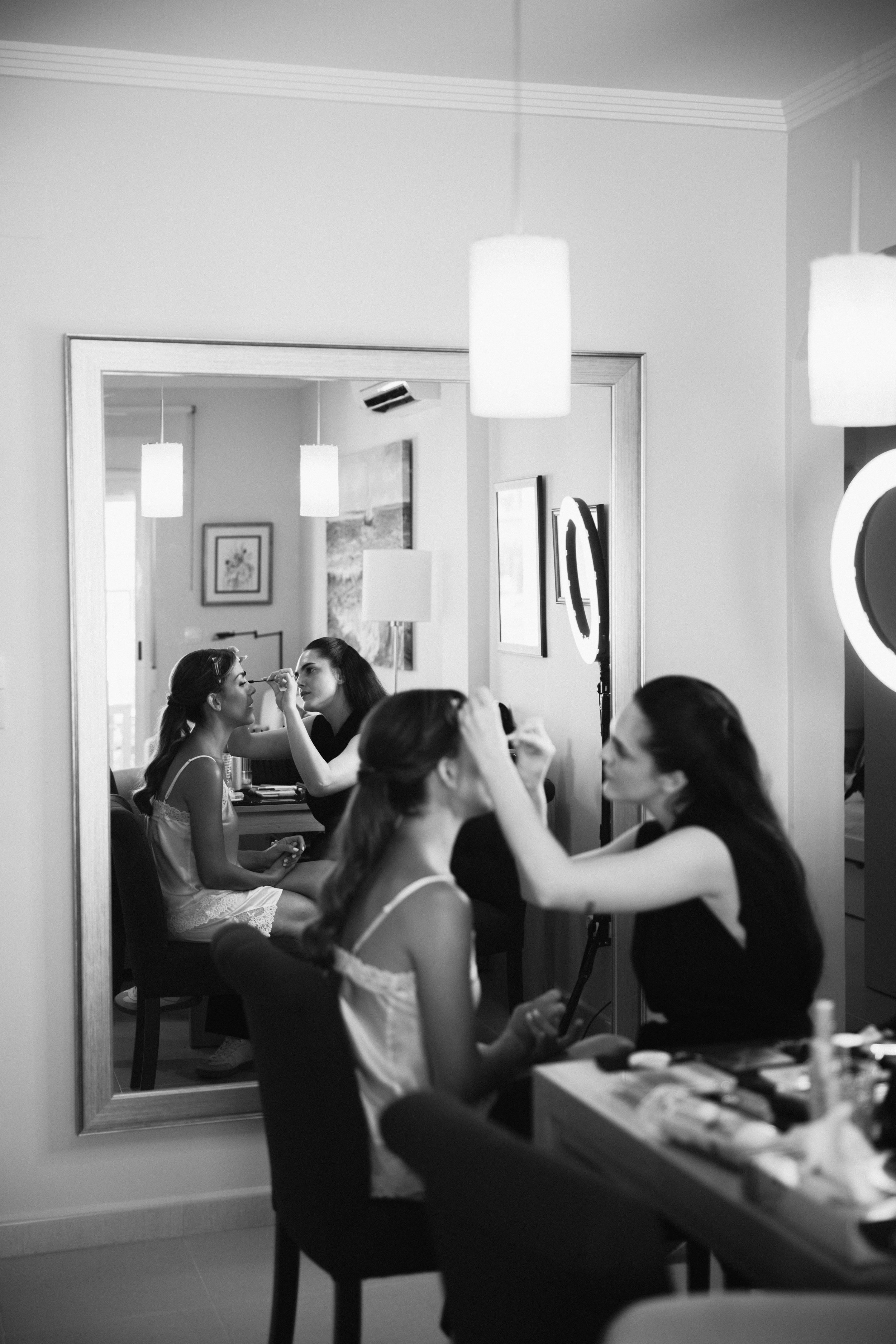 Mujeres preparando a otra para un evento frente a un espejo en una habitación de hotel o dormitorio, con maquillaje y productos de belleza sobre la mesa.