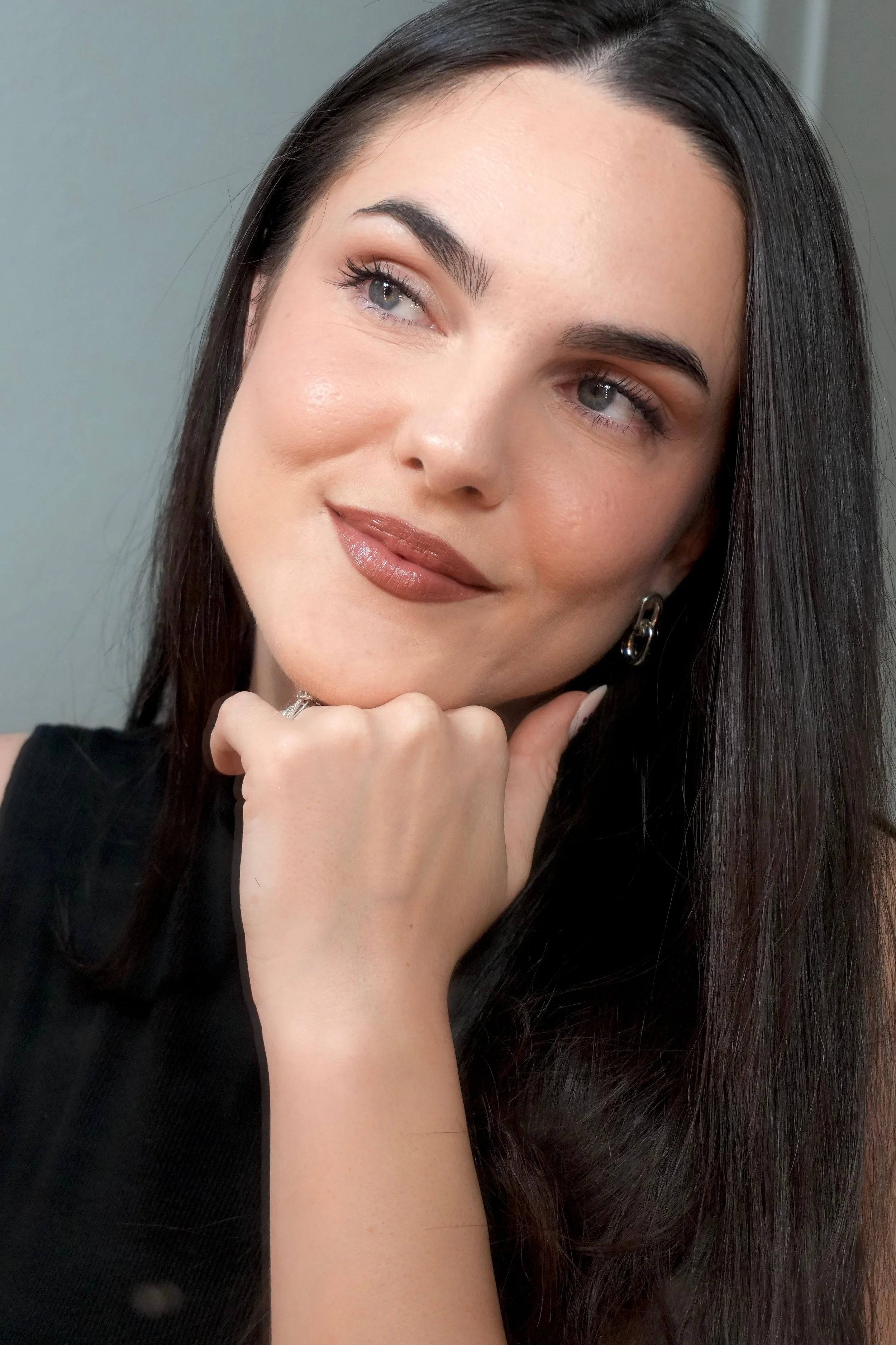 Retrato cercano de una mujer con cabello oscuro, ojos claros, maquillada, con una expresión pensativa, apoyando la mano en su rostro, usando un arete y una prenda negra.